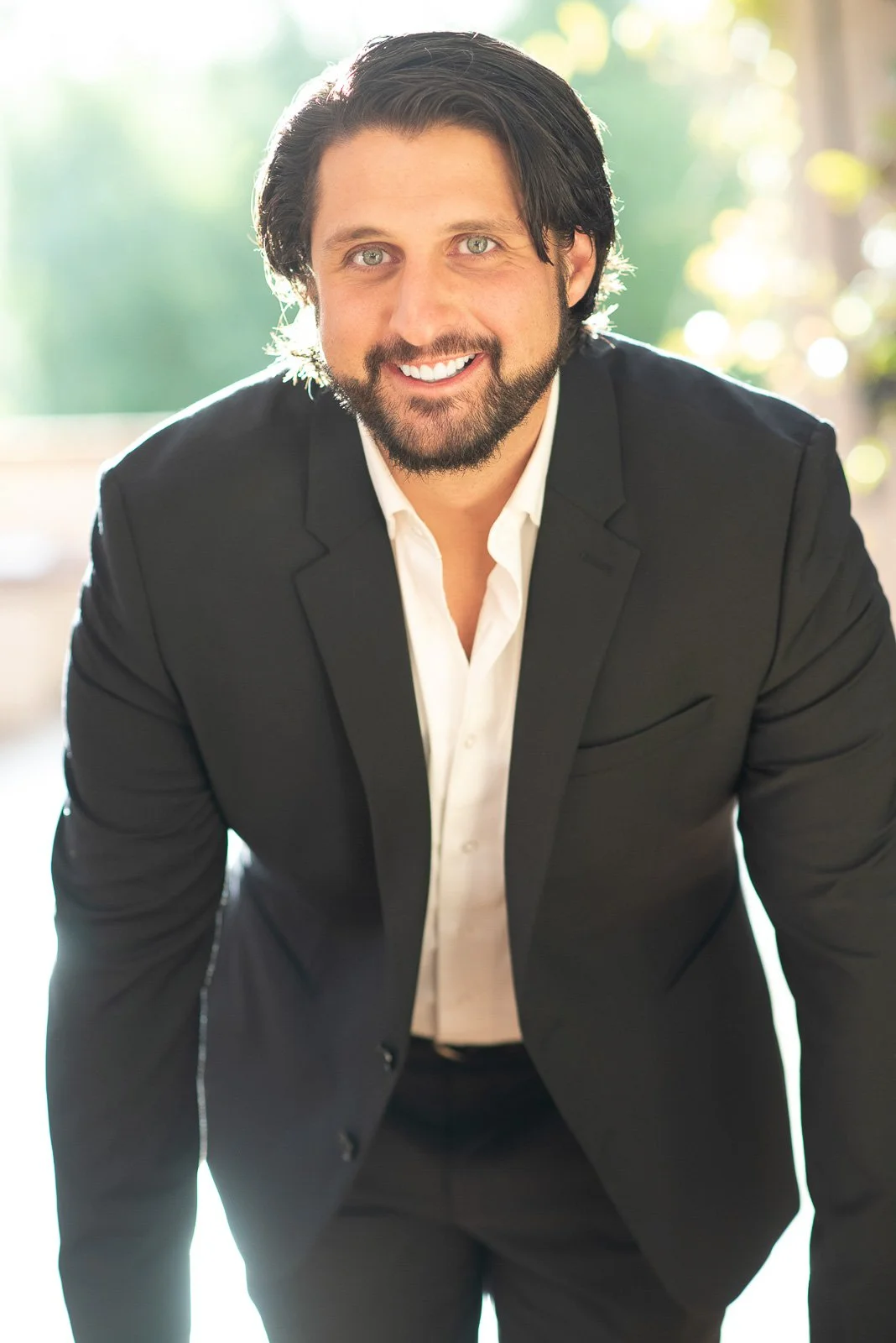 Dark haired bearded man stands with his hands on a table and sunlight behind him for his executive coaching photography session.