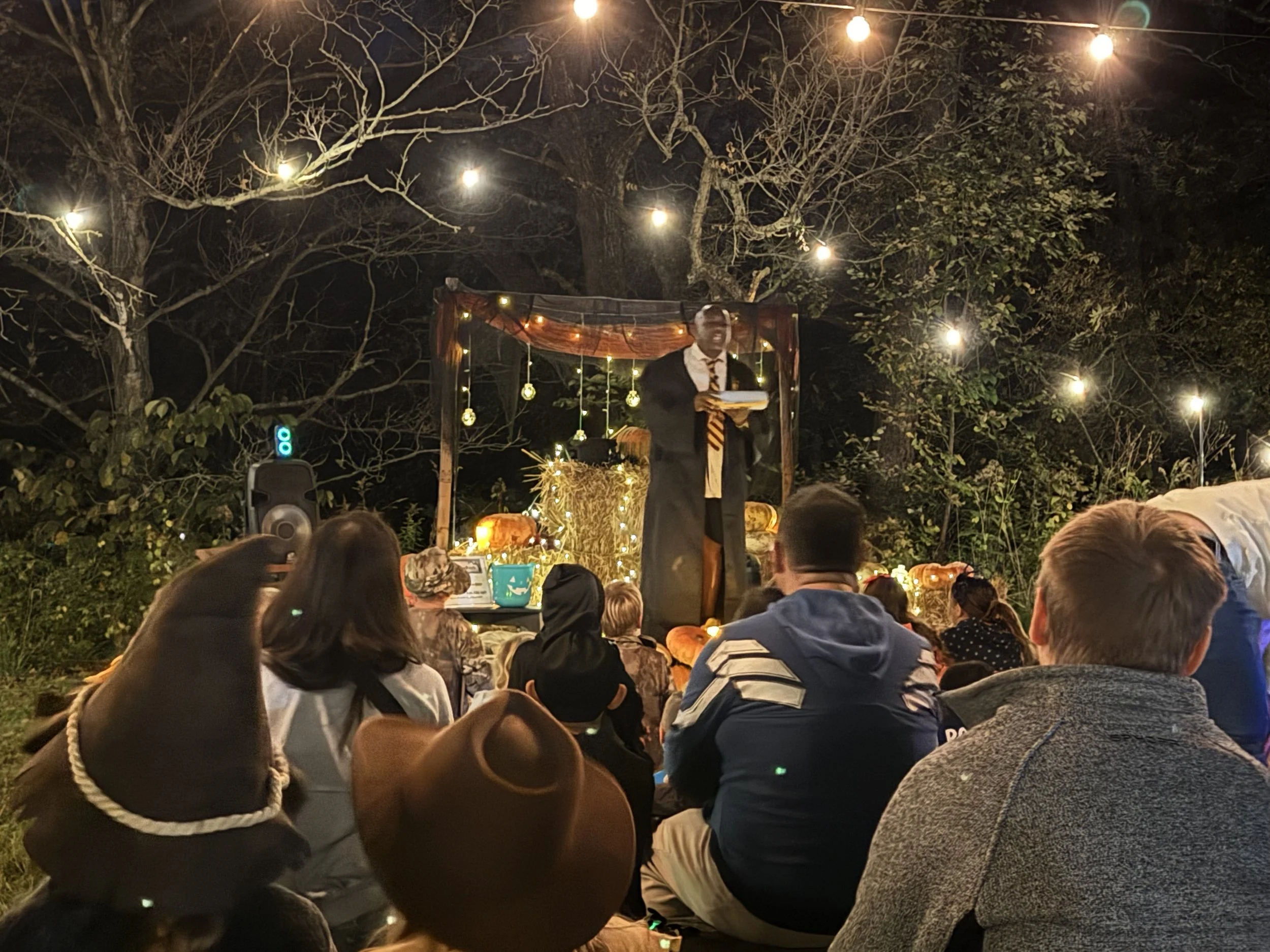 Outdoor setting with a magician on small lit stage and people in costume watching him