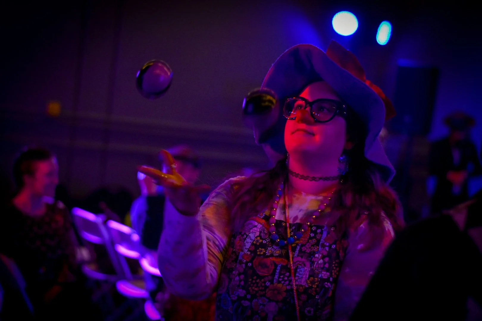 Young woman dressed as a witch with a pointed hat and glasses, juggling multiple colorful balls in a dark, colorful lighted room