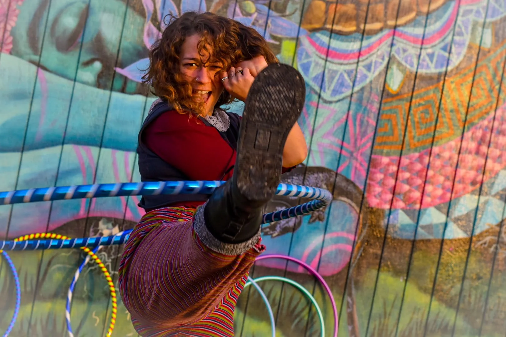 A woman with curly hair, wearing a hoodie and colorful striped pants, is smiling and putting her foot on a hula hoop while standing on a metal ring, in front of a vibrant mural with a bird and intricate designs.