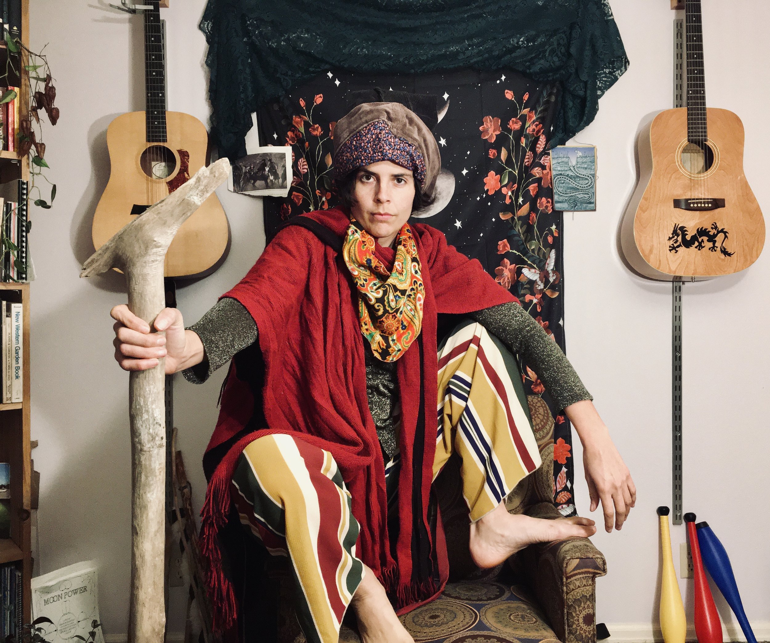 A person with dark hair, wearing a patterned hat, colorful scarf, red jacket, and striped pants, sitting on a chair with their foot on another person's knee. The person is holding a large piece of driftwood. The background has two guitars hanging on the wall, framed pictures, a bookshelf, and a colorful tapestry with floral and moon designs.