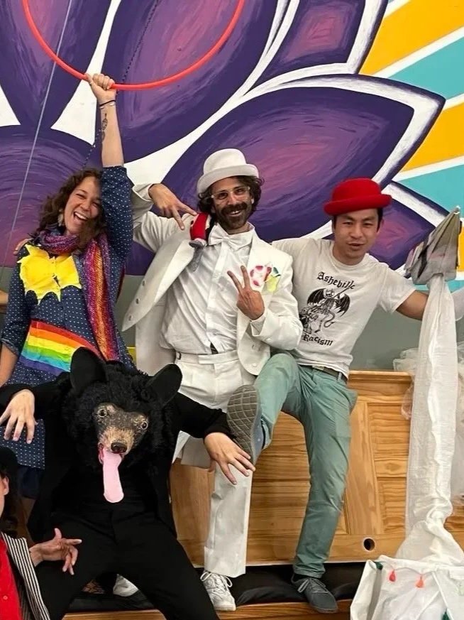 Three people in colorful, eclectic outfits; one person in a bear costume with a pink tongue, another wearing a rainbow scarf and rainbow pride flag, and the third in a red hat. They are posing in front of a vibrant purple and yellow mural.