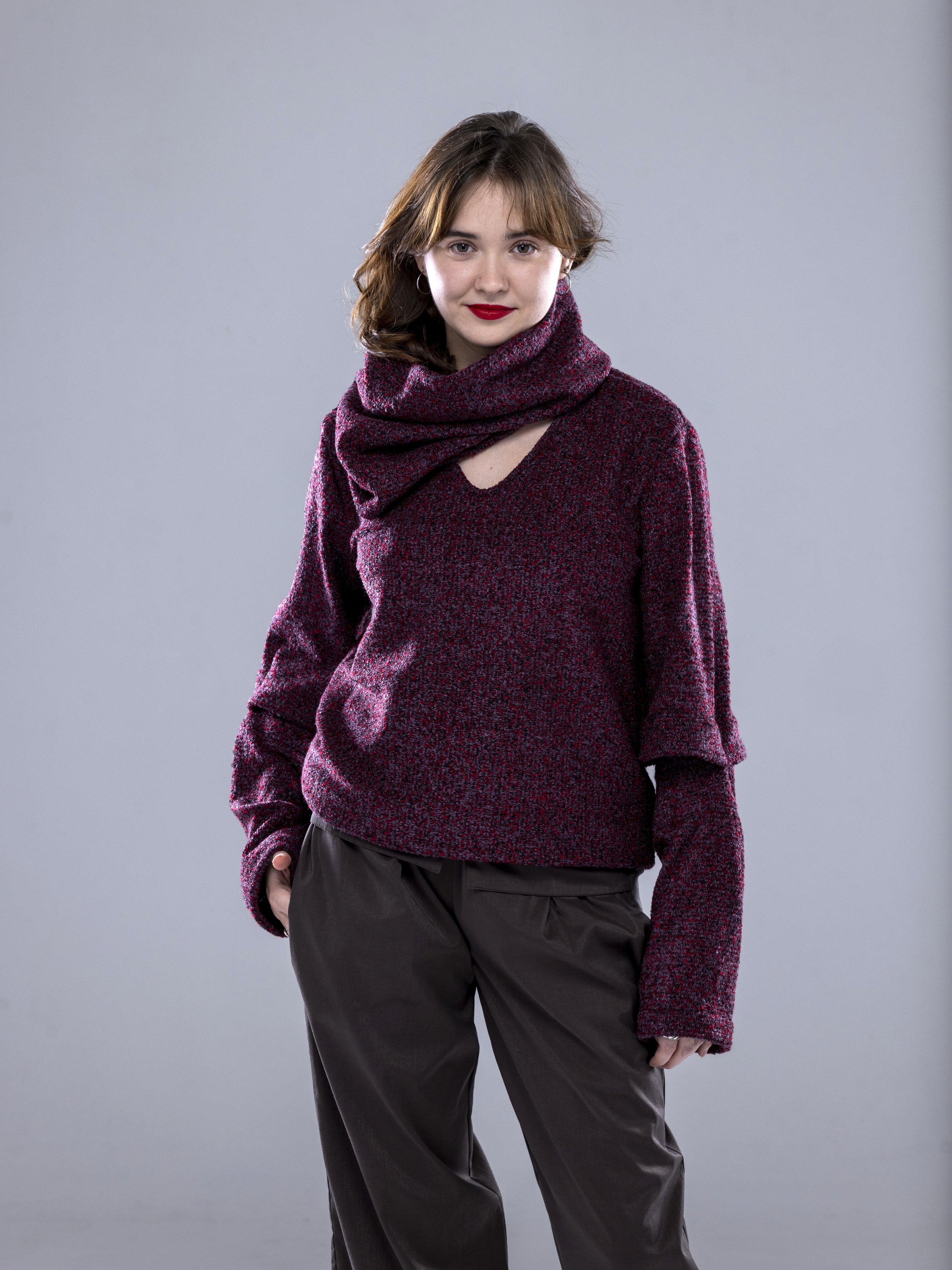 A woman with shoulder-length brown hair wearing a burgundy and black patterned sweater, black pants, and a matching scarf, standing against a plain gray background.