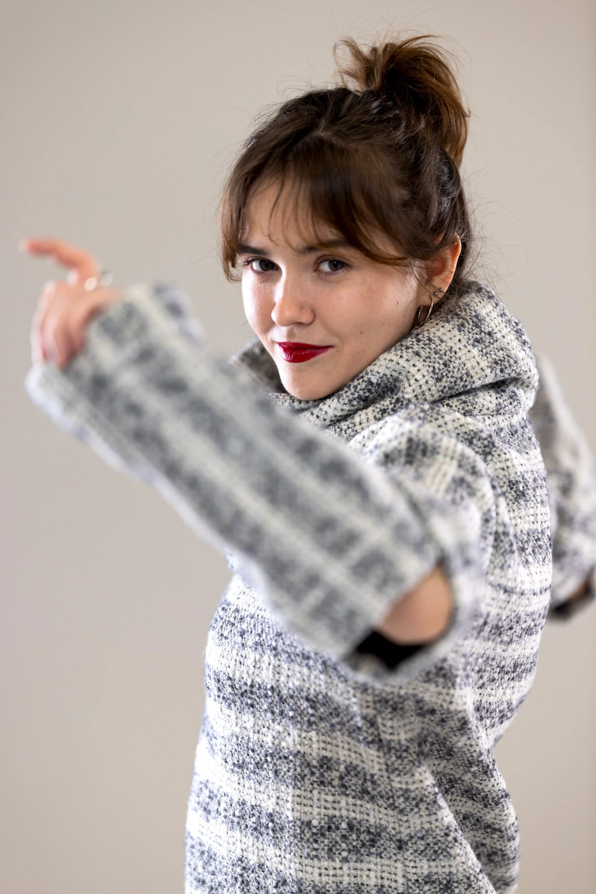 A young woman with brown hair tied in a messy bun, wearing a gray and white knitted sweater, poses with her arm extended towards the camera, looking confident with a slight smile, red lipstick, and small hoop earrings.