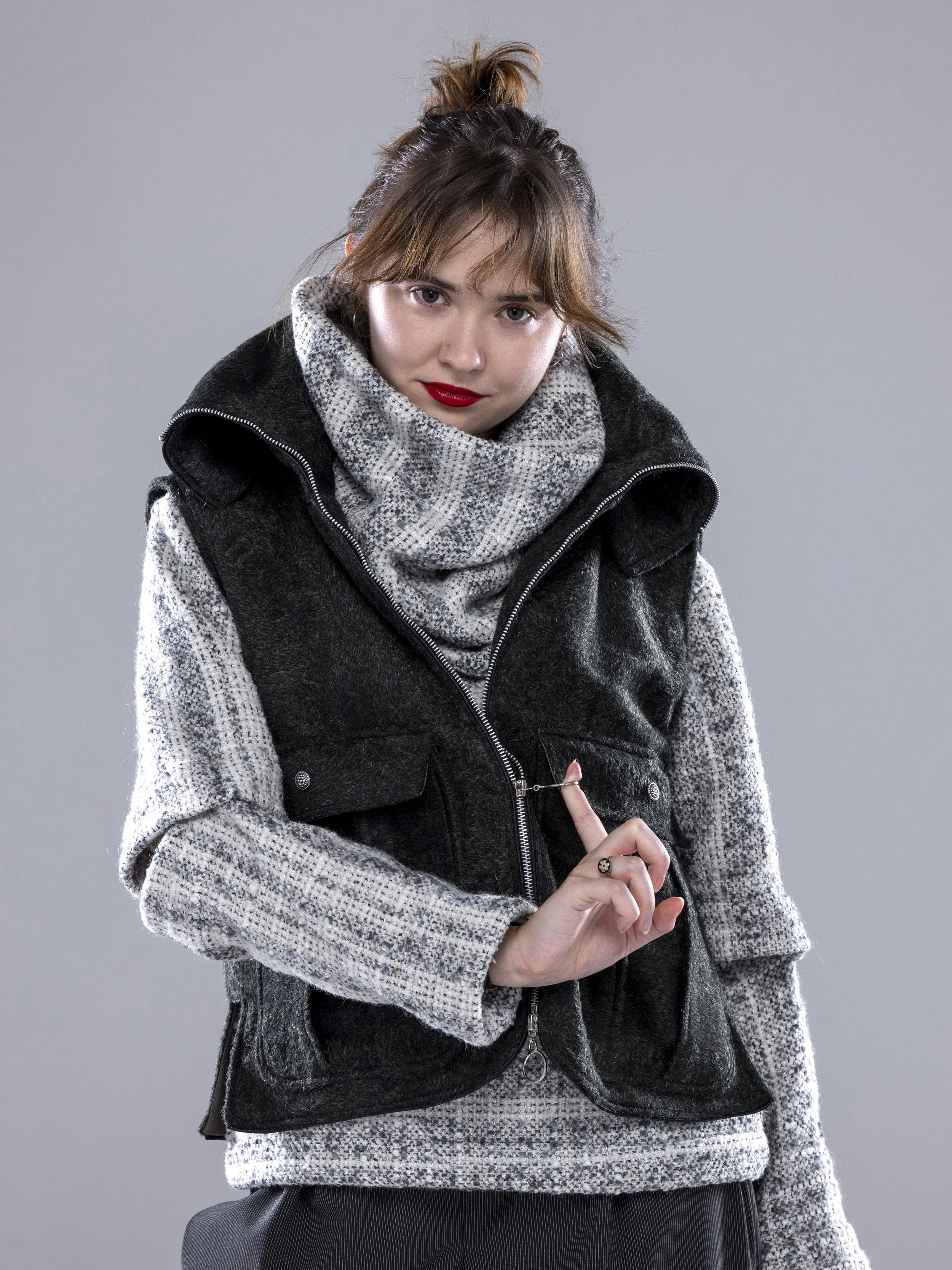 Young woman with brown hair tied up in a bun, wearing a gray scarf, black vest, and gray sweater, holding a finger up as if making a point, against a plain gray background.