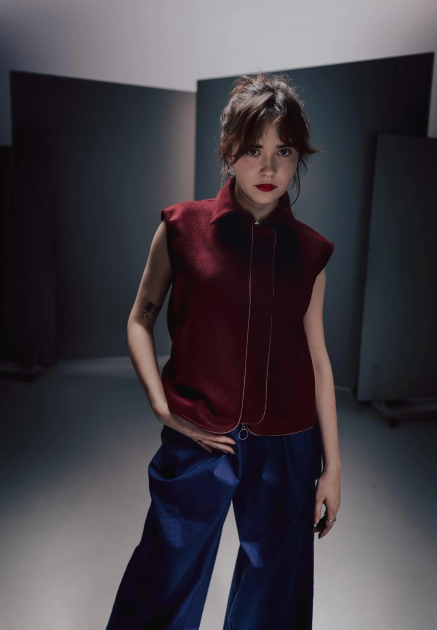 A young woman with dark hair, red lipstick, and a tattoo on her arm, standing in a dimly lit room with dark geometric panels in the background, wearing a sleeveless red vest and blue pants.