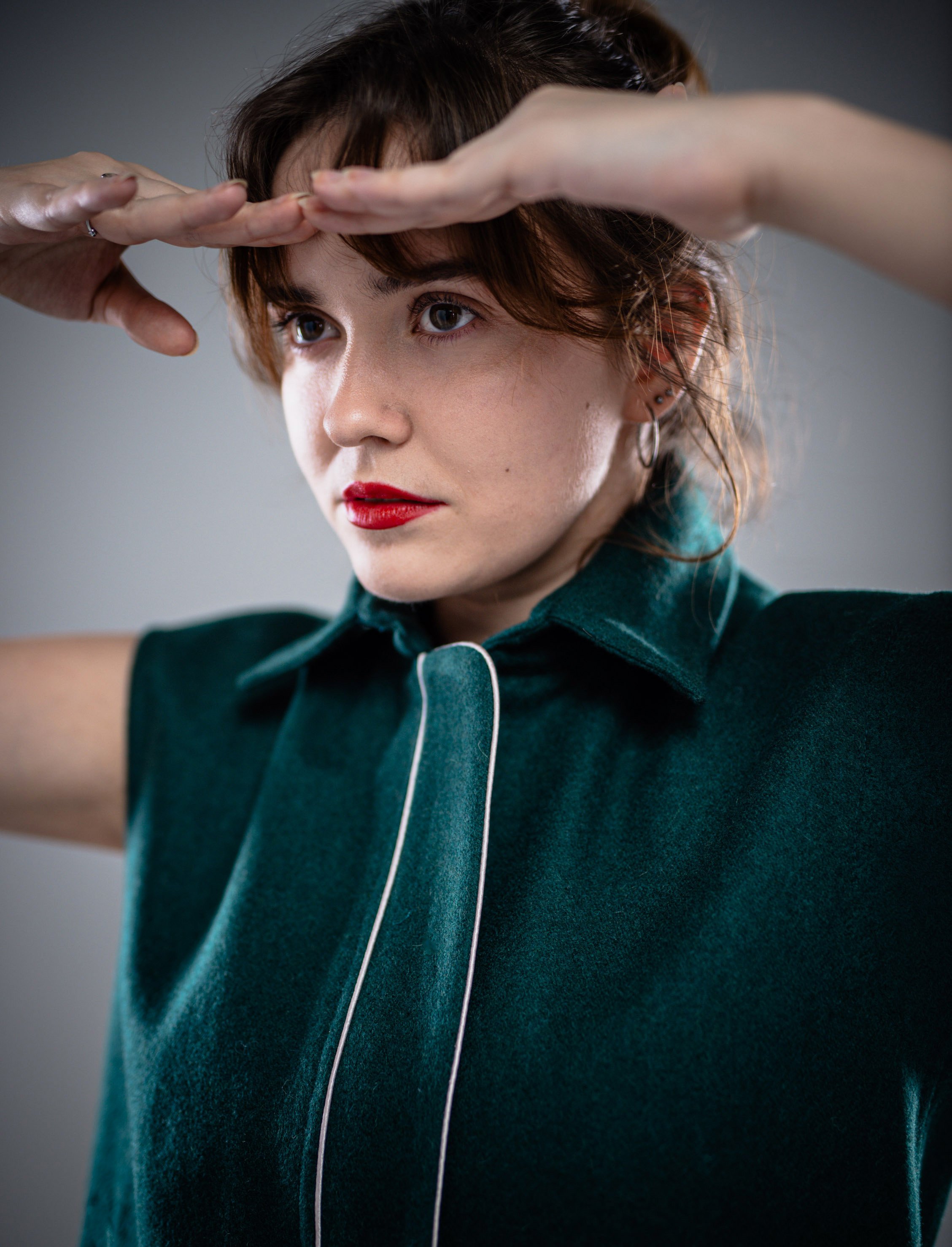 A woman with short brown hair and red lipstick wears a green sleeveless shirt with white piping, looking to the side while shielding her eyes with her hands.