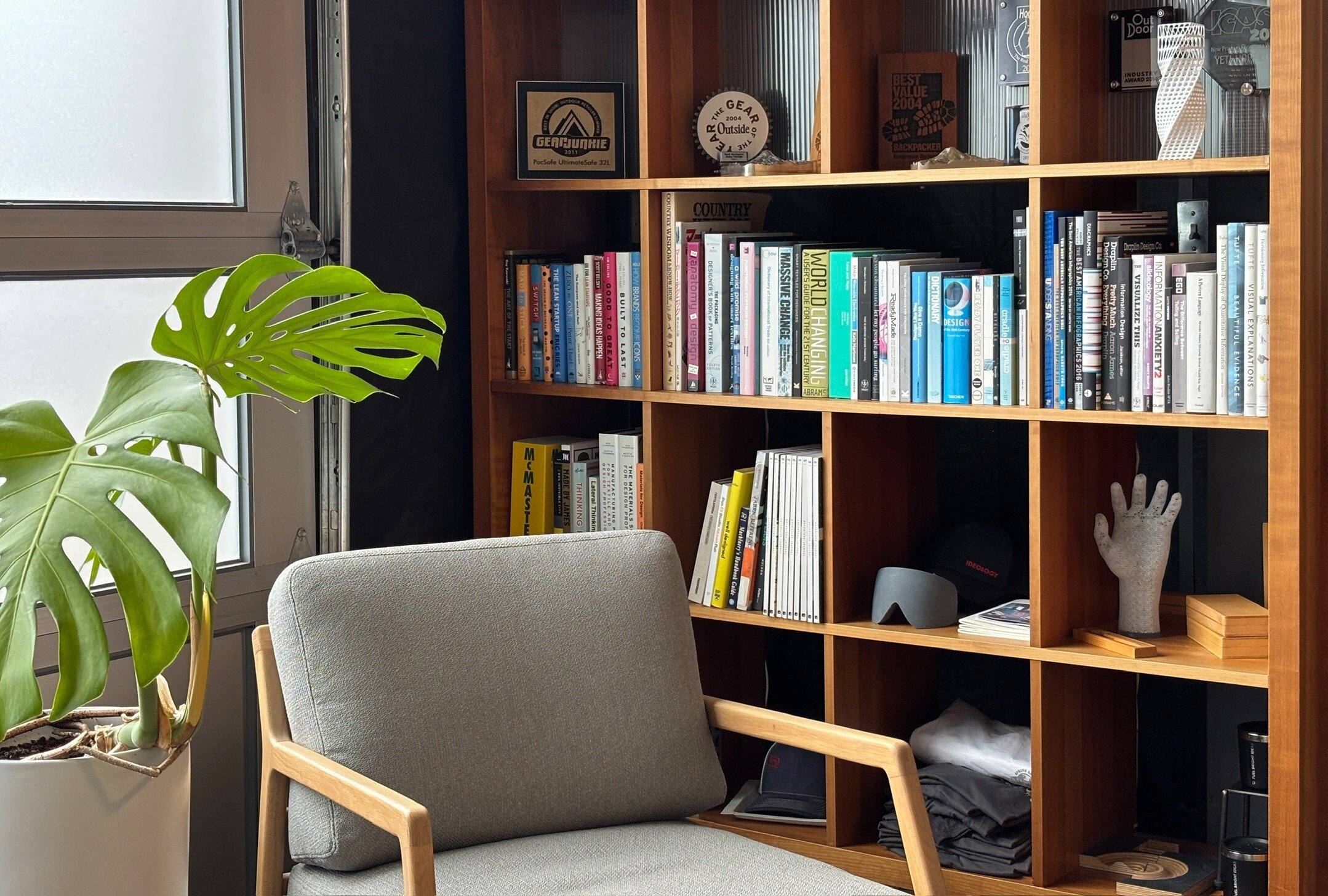 A neatly organized wooden bookshelf filled with books, medals, and decorative items in a cozy office space. A large green Monstera plant is in the foreground near a gray upholstered chair, next to a window with frosted glass.