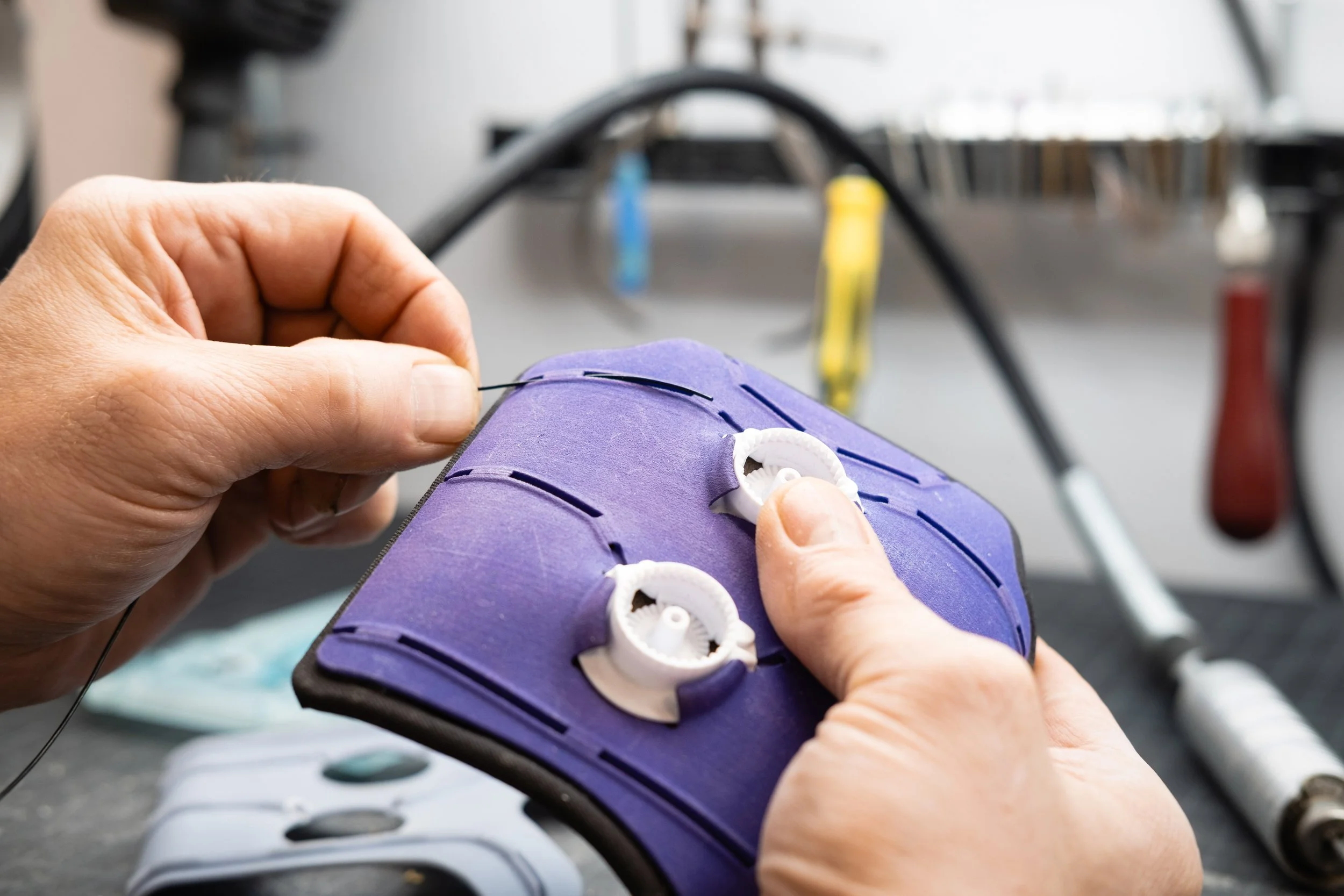 Close-up of a person's hands sewing a purple walking shoe with white gears for fitting, using a needle and thread. In the background, there are various tools and wires on a workbench.