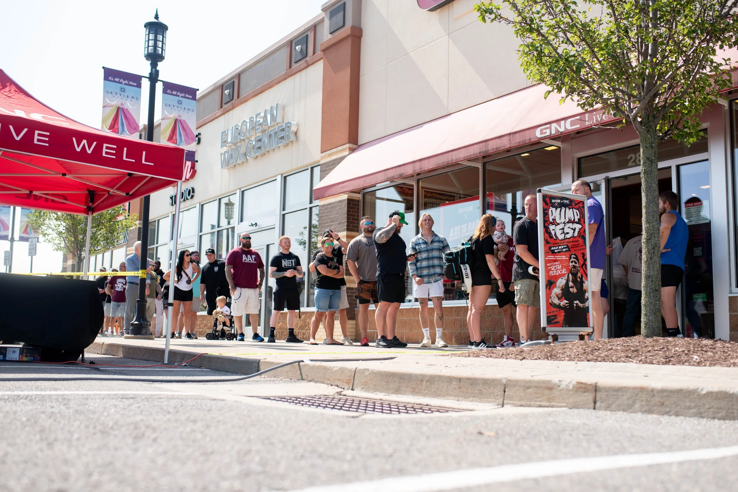 People standing in line outside a building with a sign that reads European Wax Center, on a sunny day with a tree and a red canopy nearby.