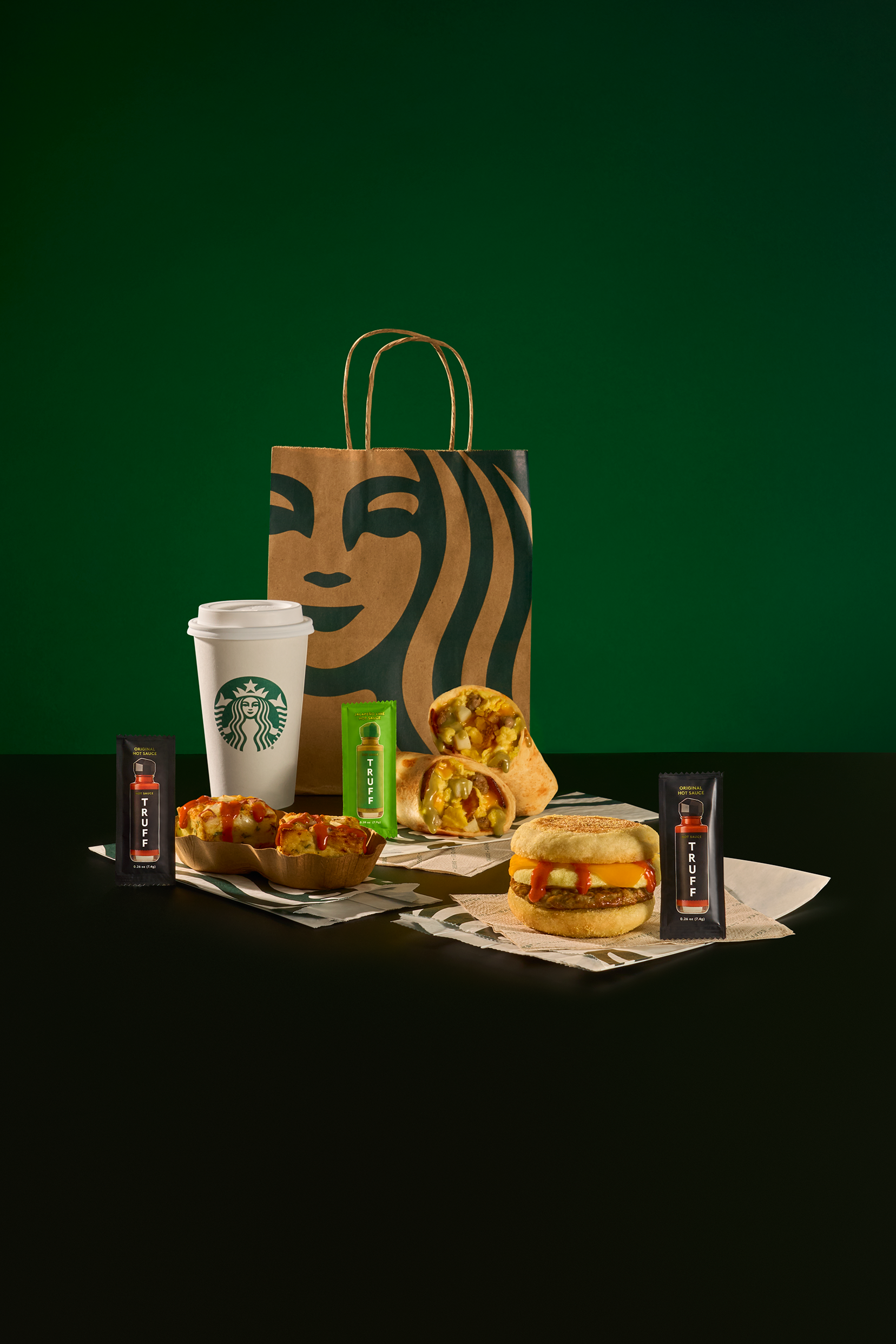 Starbucks food and drink items on a black surface with a green background, including a paper cup, a sandwich, baked food, condiments, and a paper bag with Starbucks logo.
