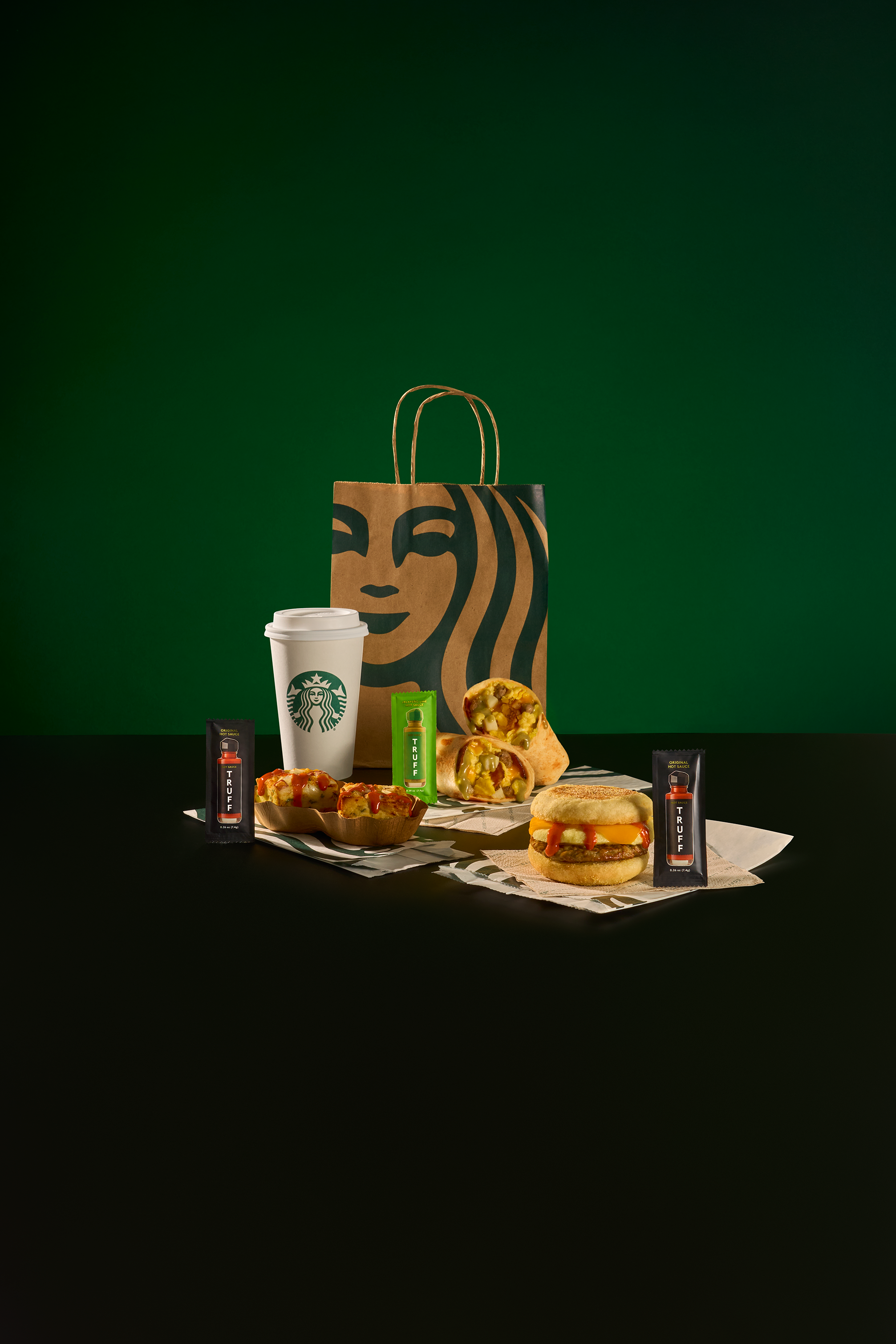 A Starbucks paper bag with a caricature of a woman with wavy hair, a white Starbucks cup, and various fast food items including sandwiches, pizza slices, and snack foods, all on a black surface with a green background.