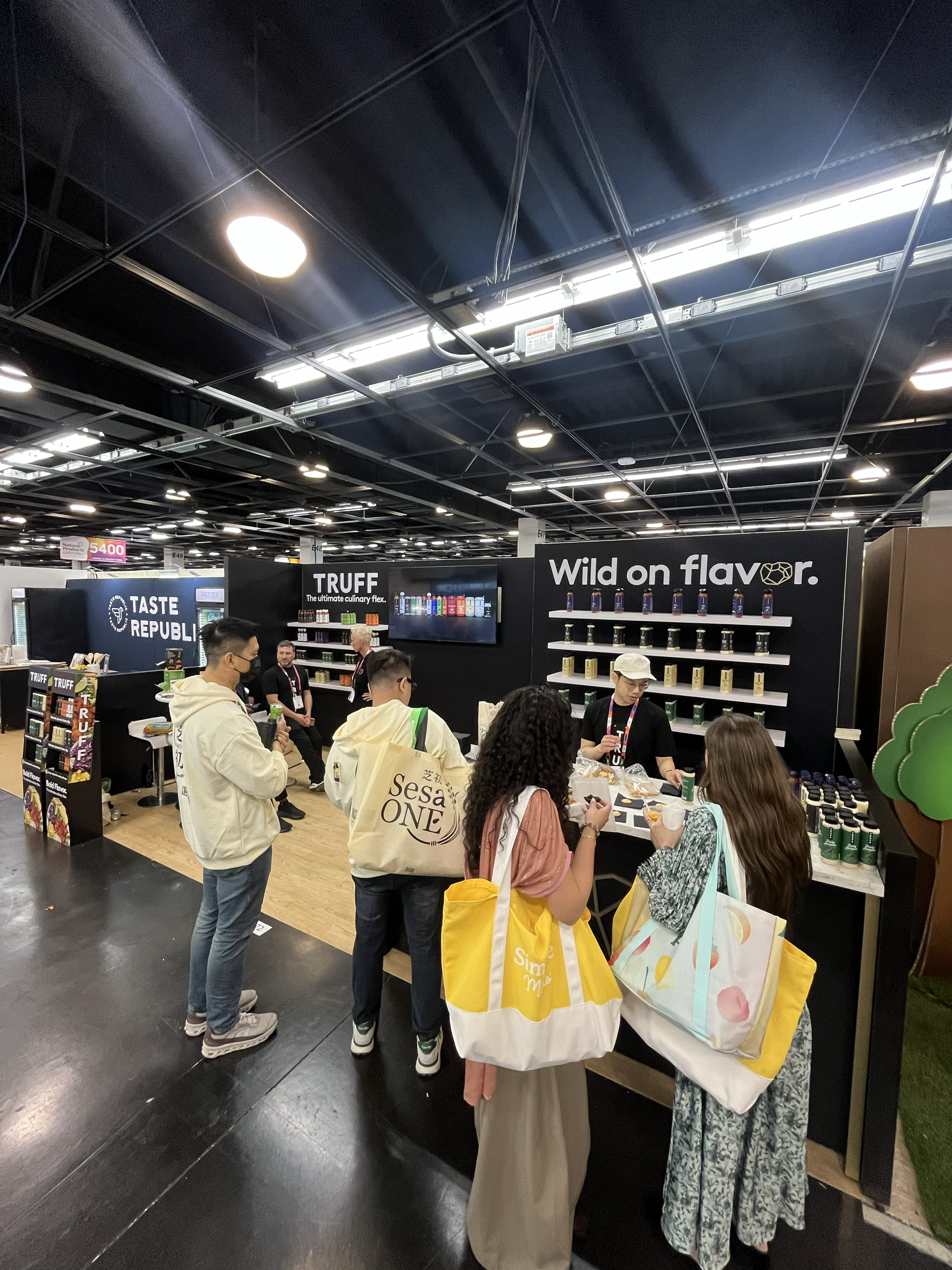 People shopping and browsing at a trade show or convention with display booths and products on shelves, including a booth with the text "Wild on flavor" and others showcasing various items.
