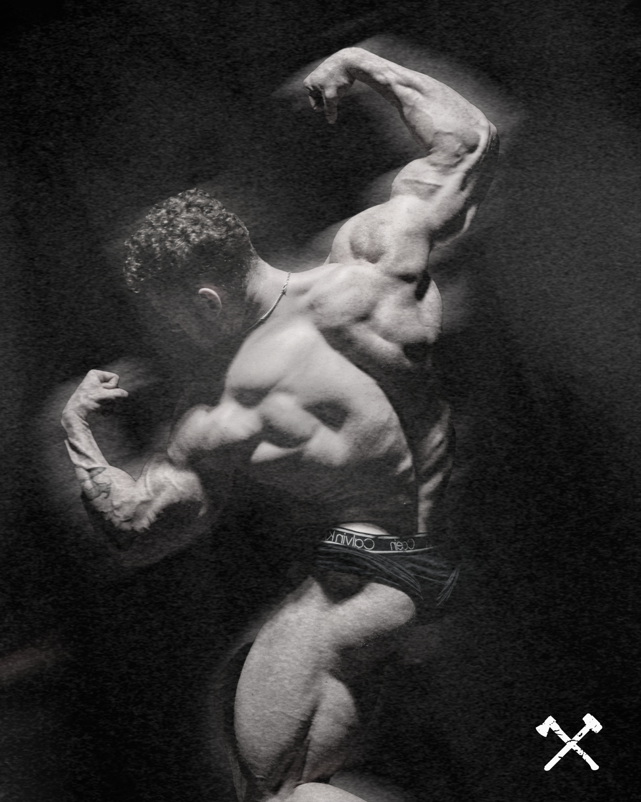 A black and white photo of a shirtless man flexing his bicep with a large muscular physique, looking down. He is wearing Calvins underwear and is in a dark background with a watermark of cutlery in the bottom right corner.