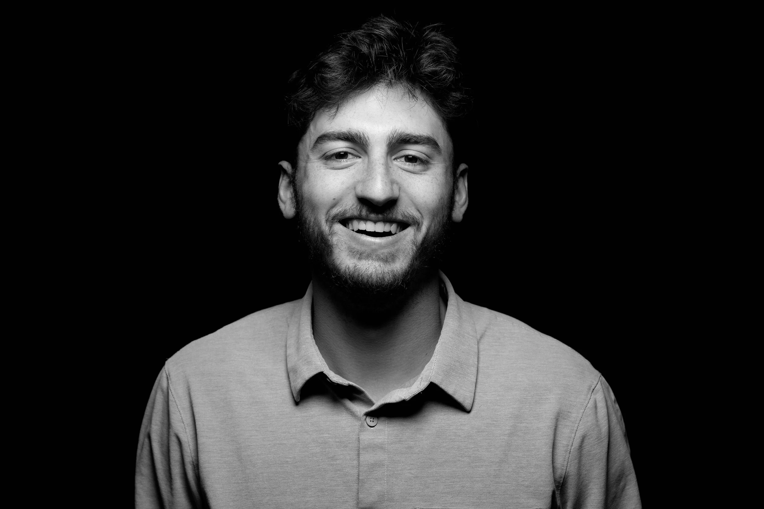 Black and white portrait of a young man with a beard smiling against a black background.