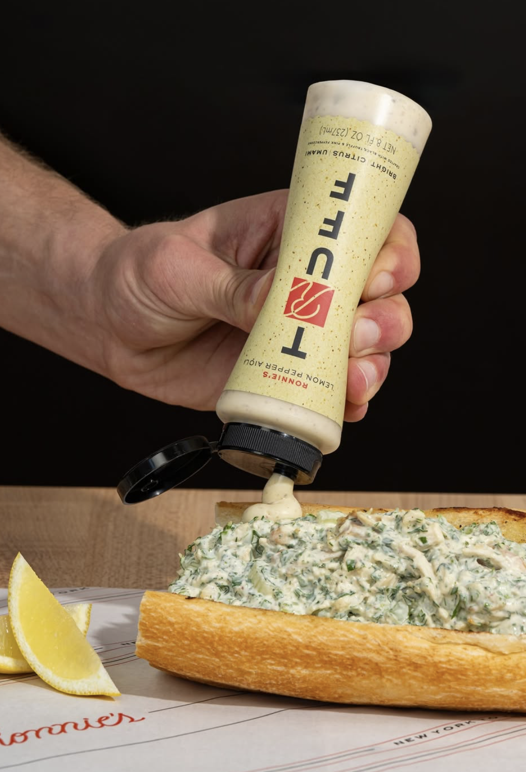 Person squeezing a tube of mayonnaise onto a seafood roll sandwich, with lemon wedges on the side.