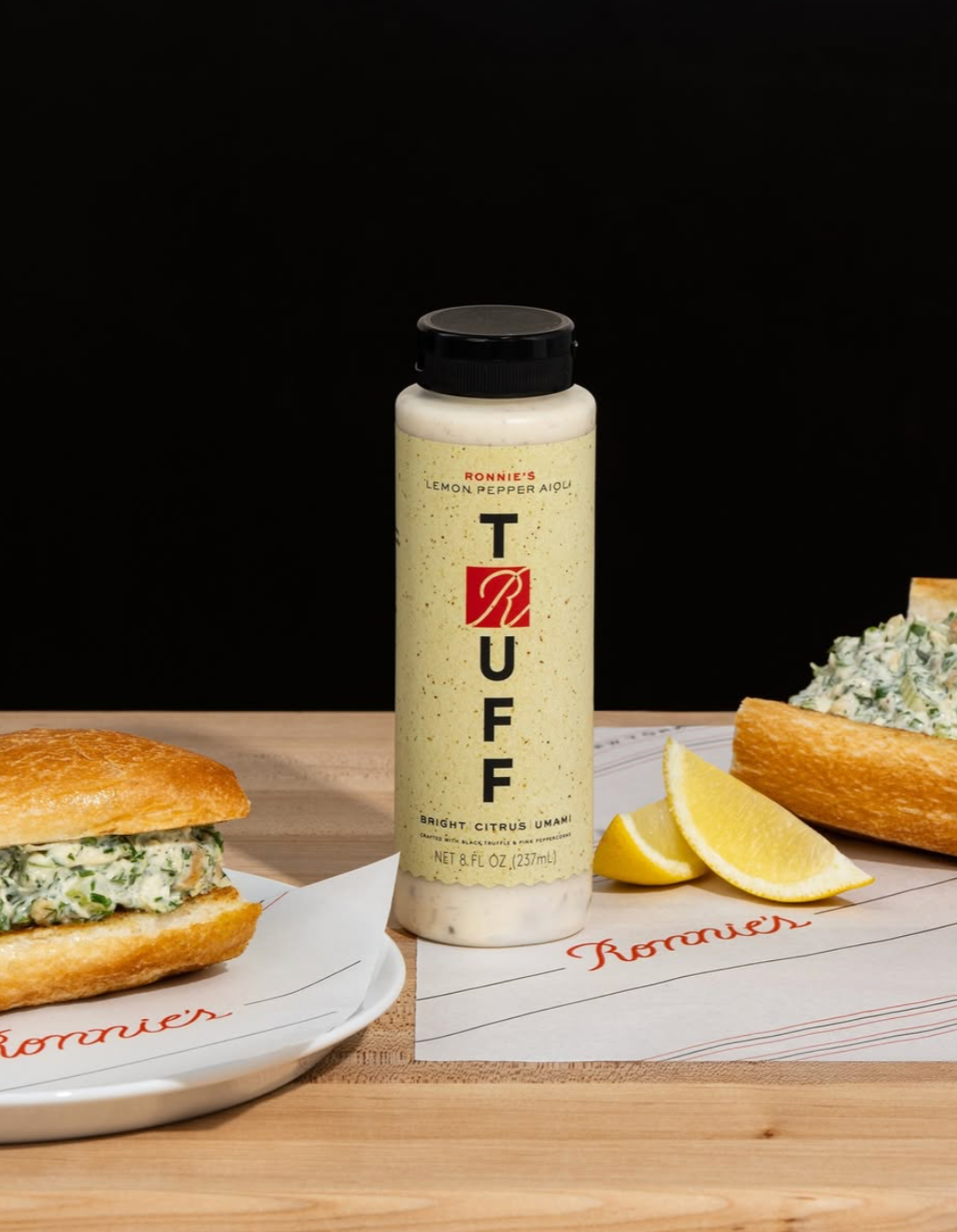 A bottle of Ronie's Lemon Pepper Tuff sauce on a wooden surface with two sandwiches, lemon wedges, and a piece of bread with a creamy herb mixture, against a black background.