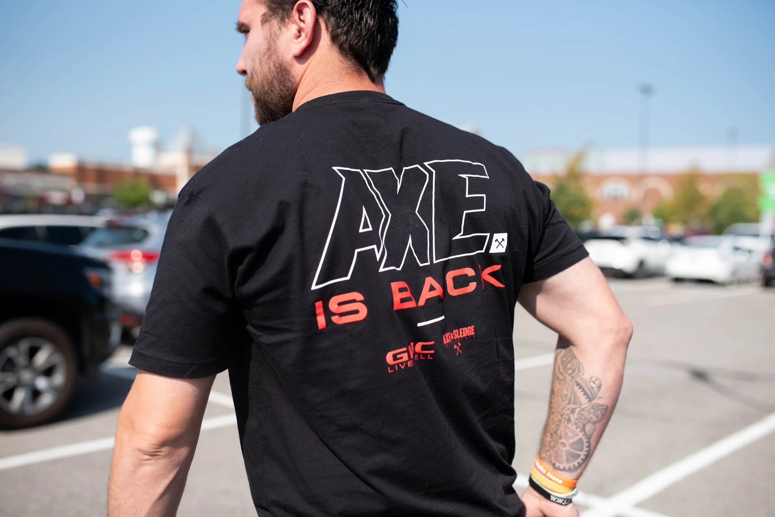 A man wearing a black T-shirt with the text 'AXE IS BACK' on the back, standing in a parking lot with cars and buildings in the background.
