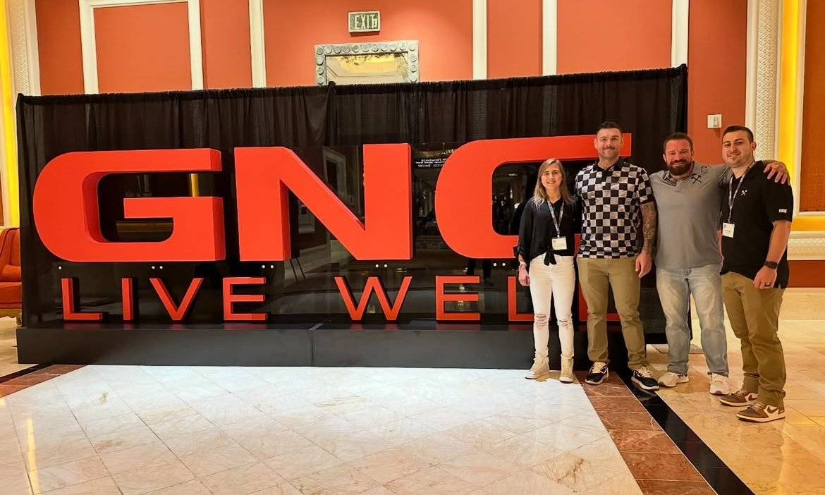 Four people standing in front of a large sign that reads "GNC LIVE WELL" at an indoor event or conference.