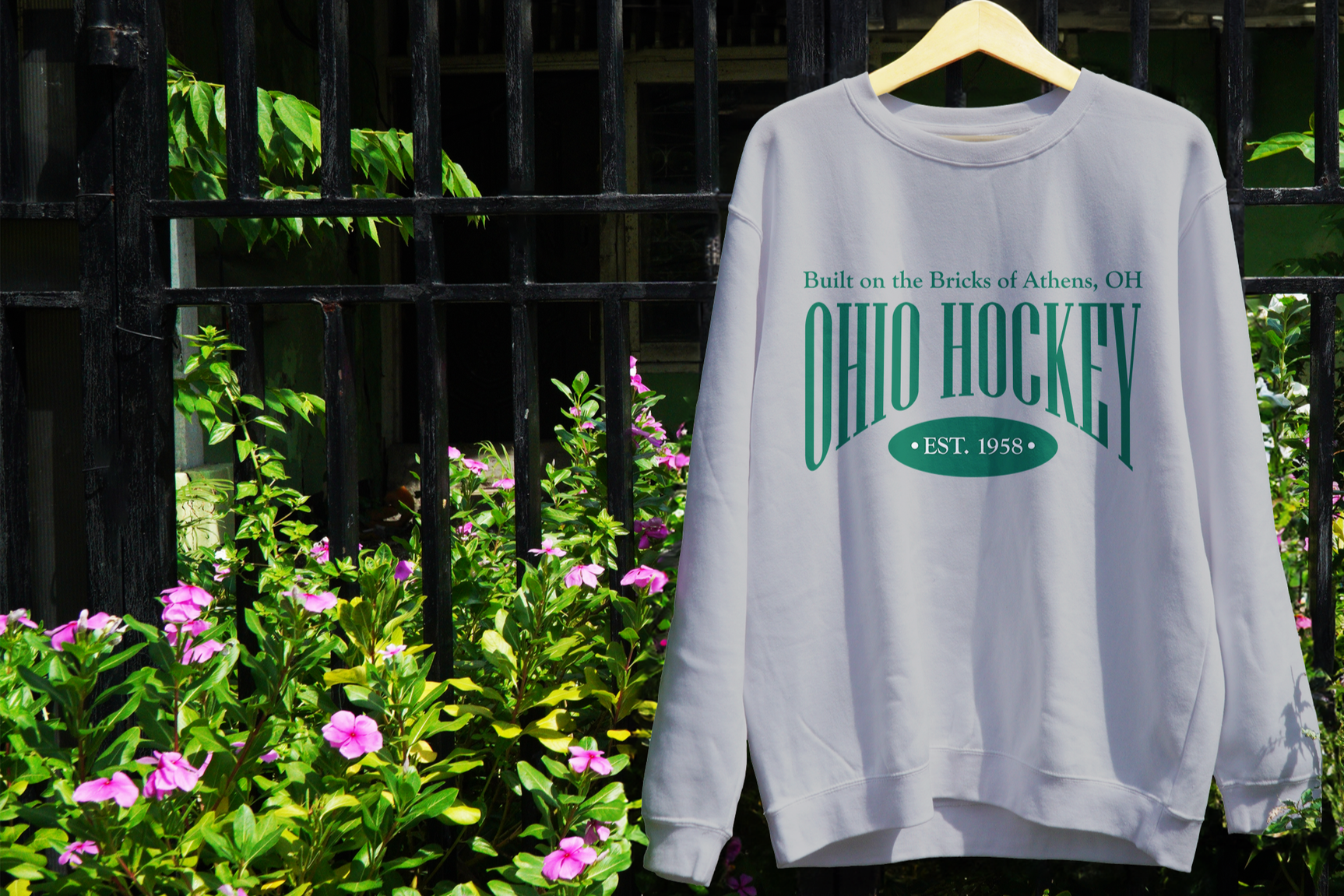 A white sweatshirt hanging on a wooden hanger against a black metal fence with pink flowers and green foliage in the background. The sweatshirt has green text that reads 'Built on the Bricks of Athens, OH,' 'OHIO HOCKEY,' and 'EST. 1958.'