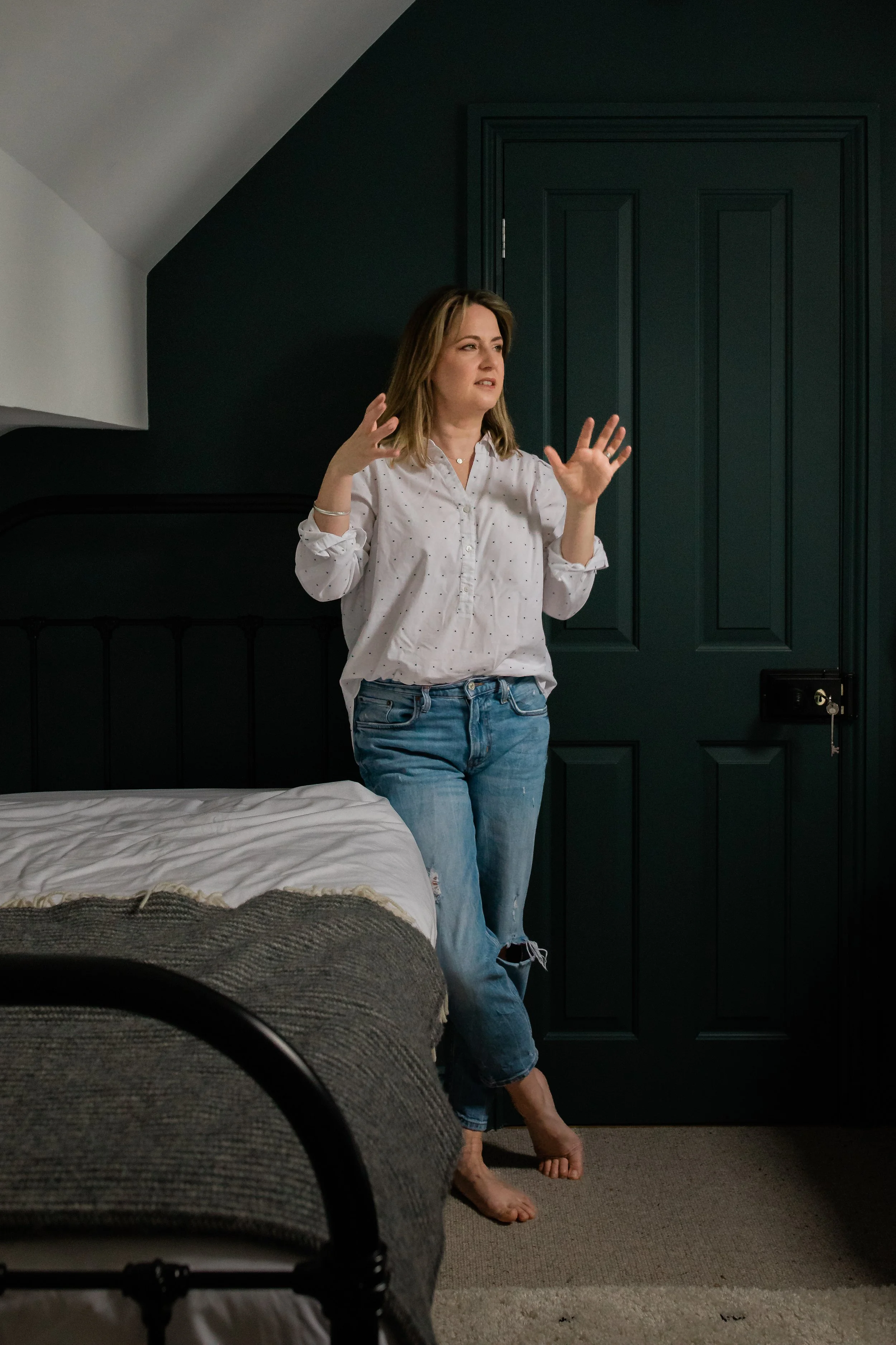 A woman with shoulder-length hair, wearing a white blouse and ripped jeans, standing barefoot in a bedroom near a dark green door, gesturing with her hands.