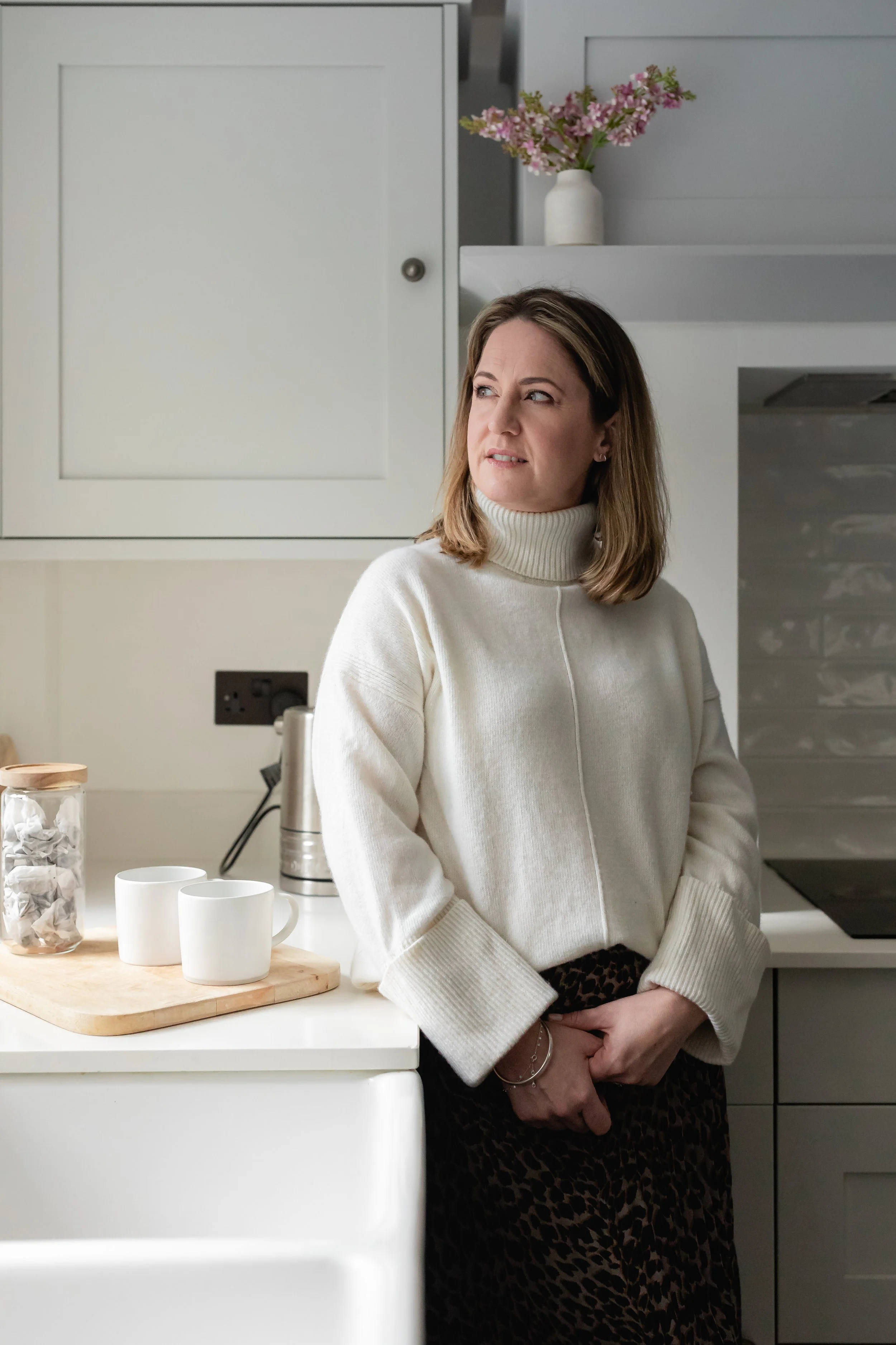 Katherine Scott of Katherine Scott Coaching with shoulder-length brown hair, wearing a green blouse and ripped jeans, smiling and leaning on a gray sofa in a bright living room with a window, a white cabinet, and some decorative plants.