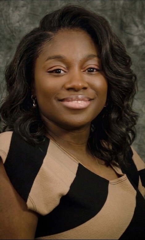 A portrait of a smiling African American woman with shoulder-length dark hair, wearing a beige and black patterned dress, gold earrings, and a delicate gold necklace, against a dark textured background.