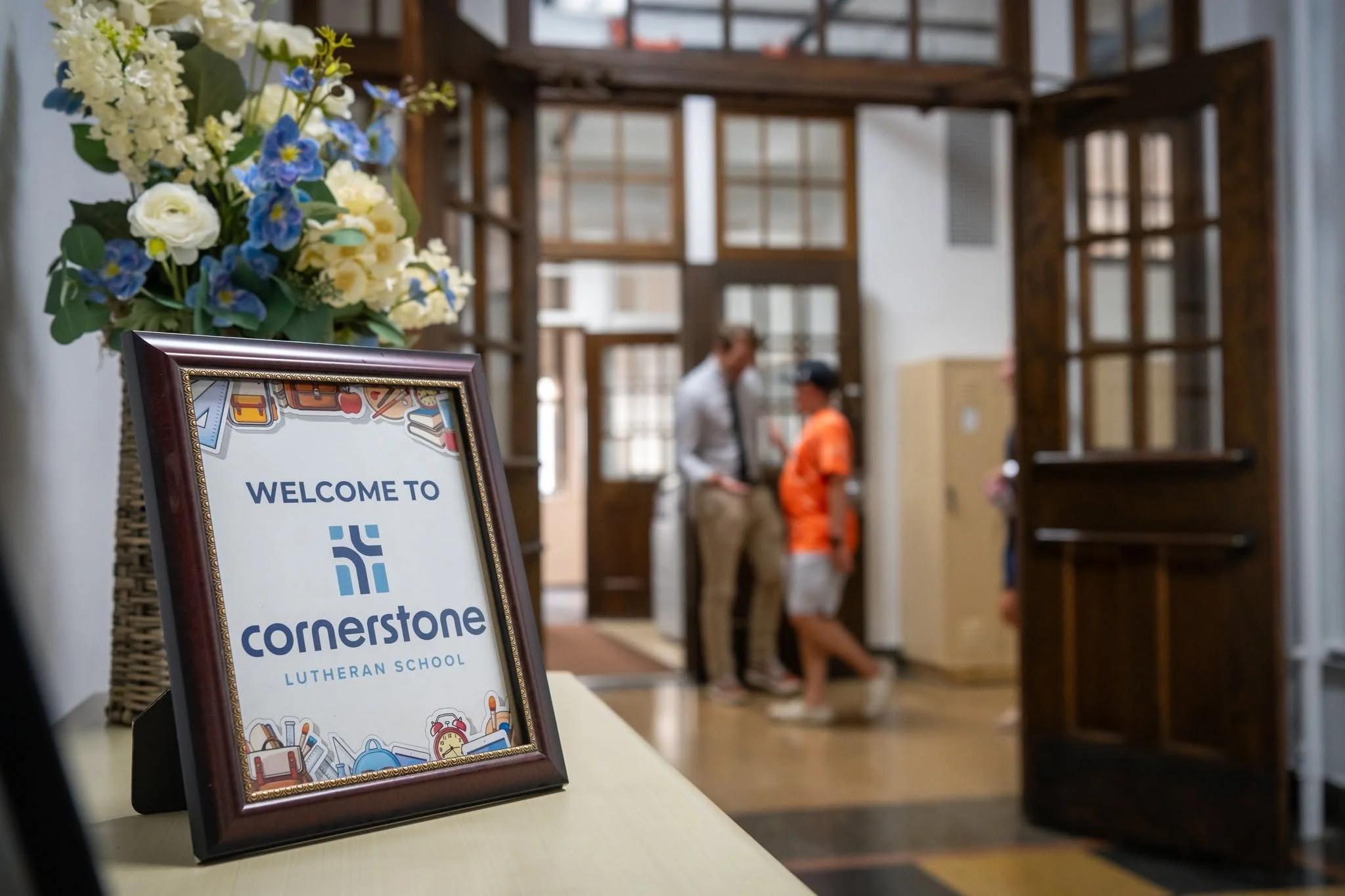 Welcome to Cornerstone Lutheran School sign sits on a desk with two figures in the background