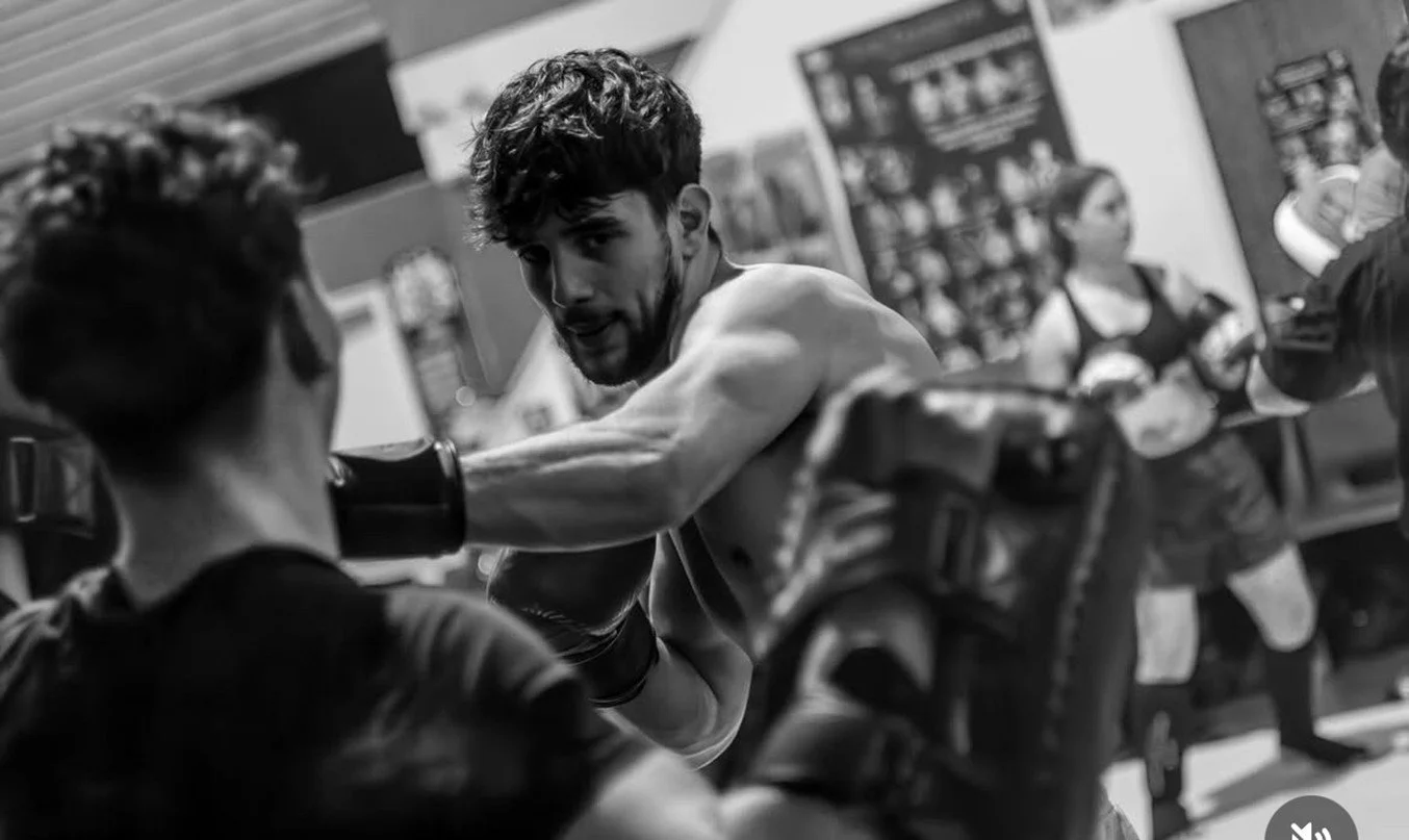 Black and white photo of a male boxer throwing a punch at a training partner in a gym, with a woman and other people in the background.