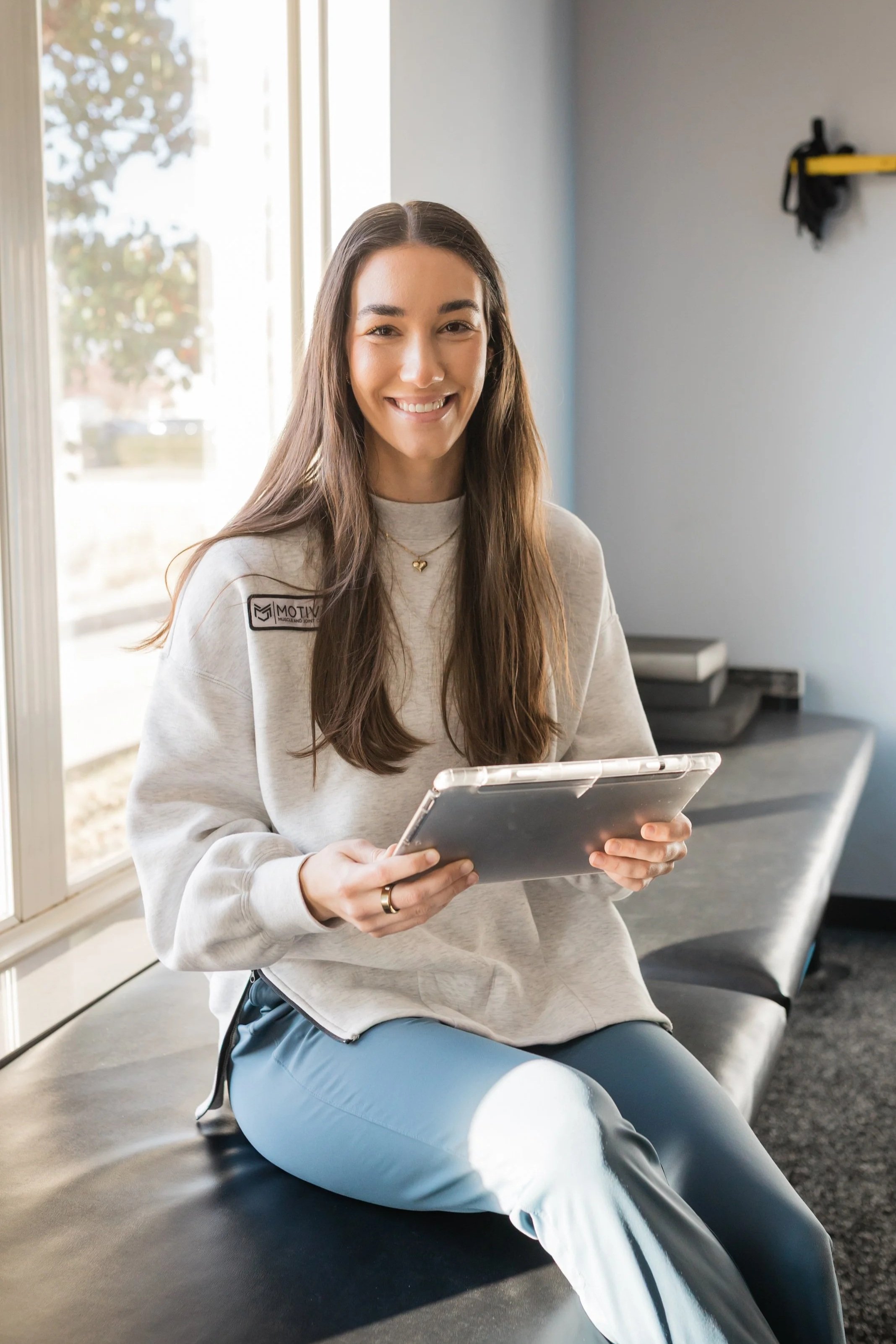 Young woman sitting on a bench by a large window, smiling and holding a tablet, wearing a light gray sweatshirt and light blue pants.
