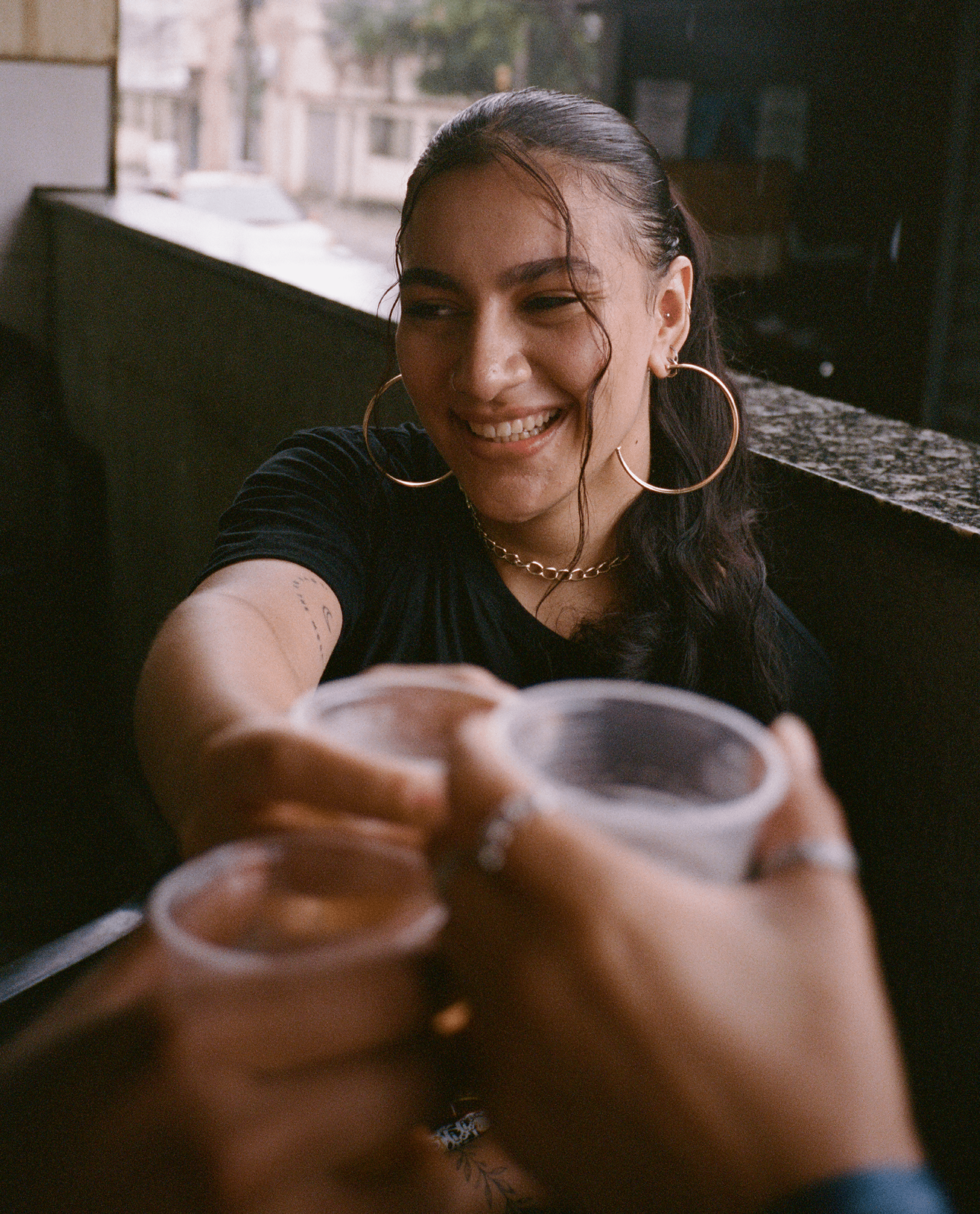A woman with dark hair and large hoop earrings smiling while reaching out for a drink in a plastic cup from a group of friends.