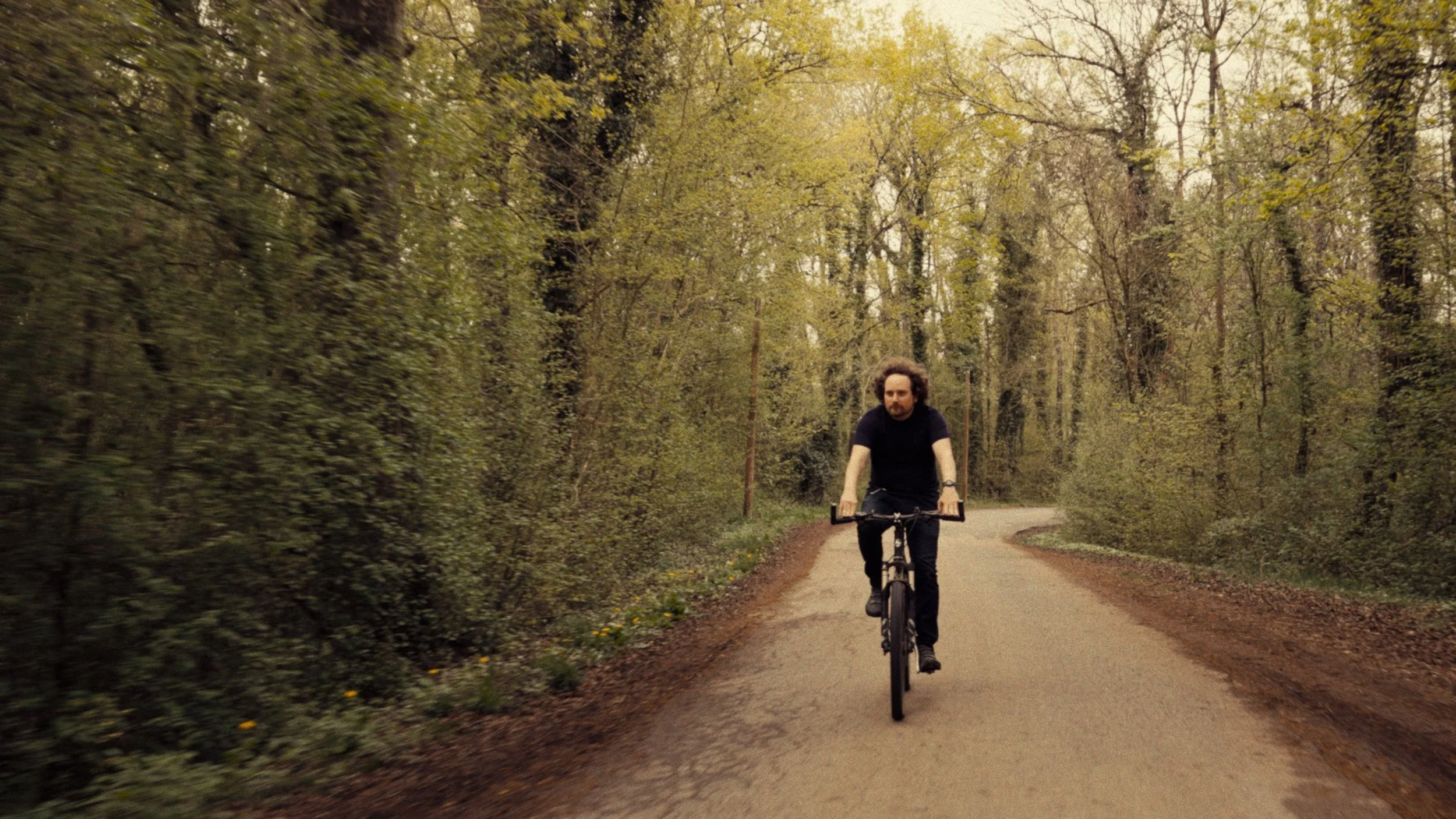 A man with curly hair riding a bicycle on a winding forest path surrounded by trees with budding leaves.