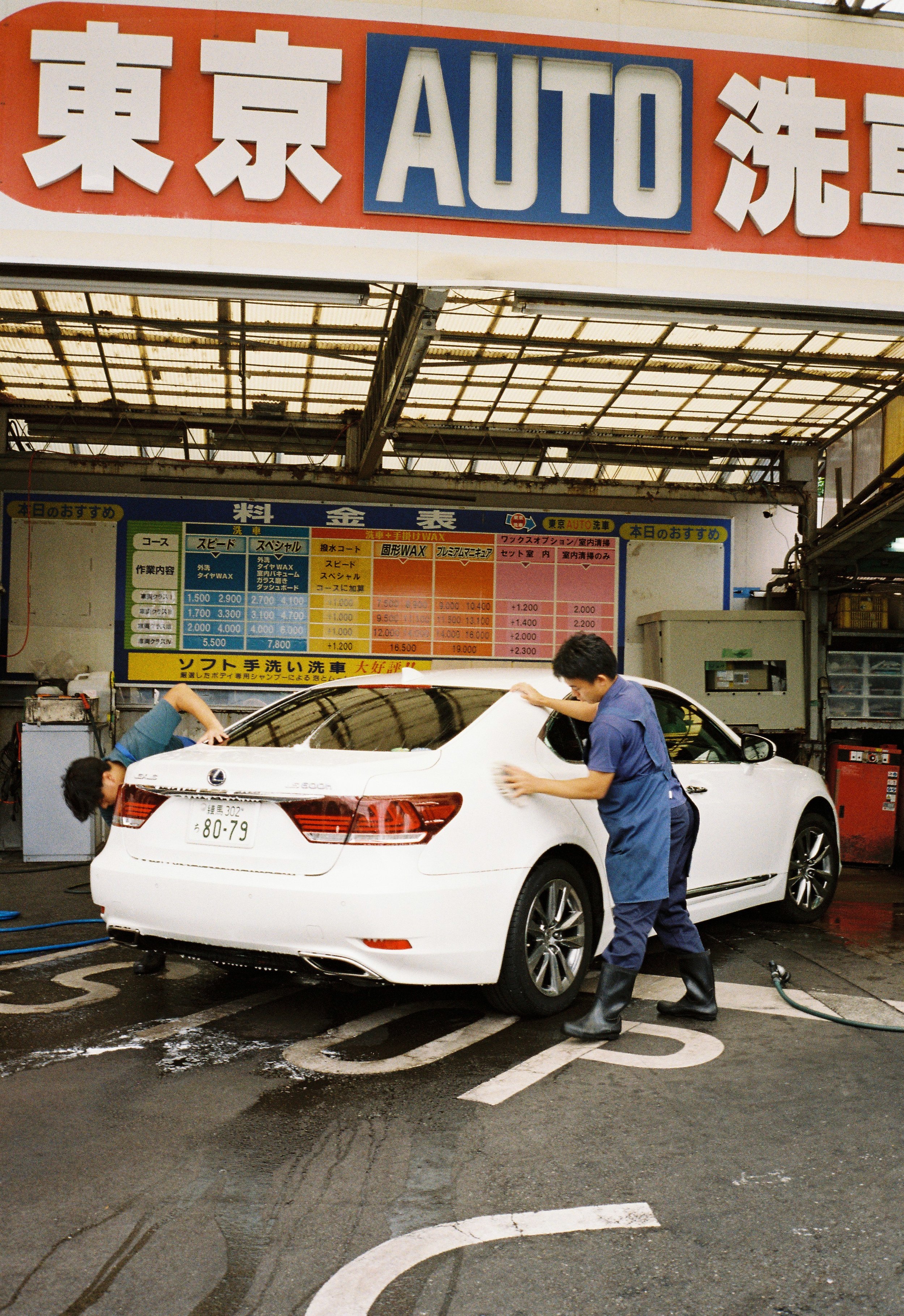 Two men washing a white Lexus car at an outdoor car wash station with a sign in Japanese and the word 'AUTO' above.