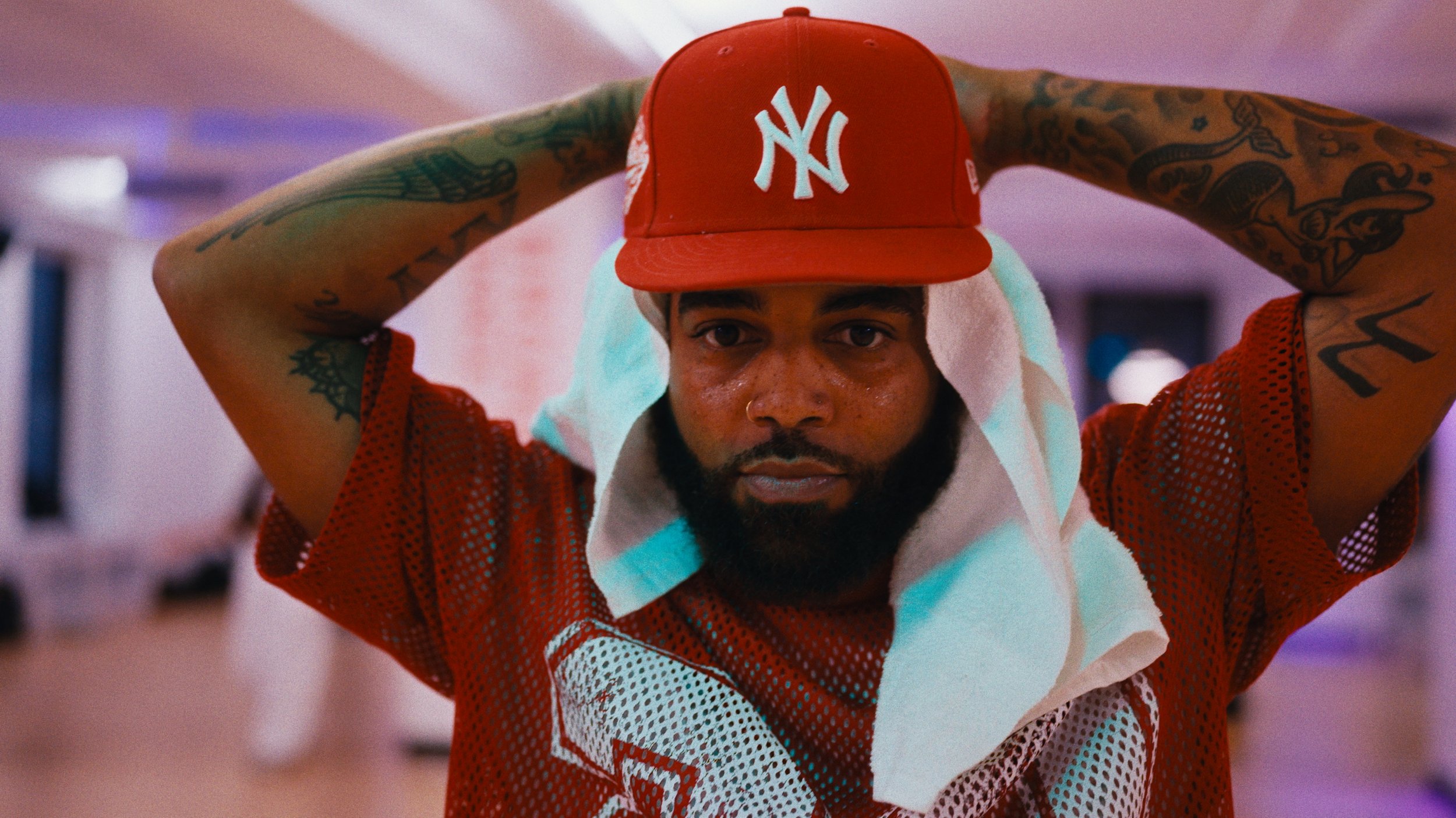 A man with a beard, tattoos on arms, and sweat on face, wearing a red New York Yankees cap and a red mesh sports jersey, with a white towel draped over his neck, in an indoor gym or fitness setting.