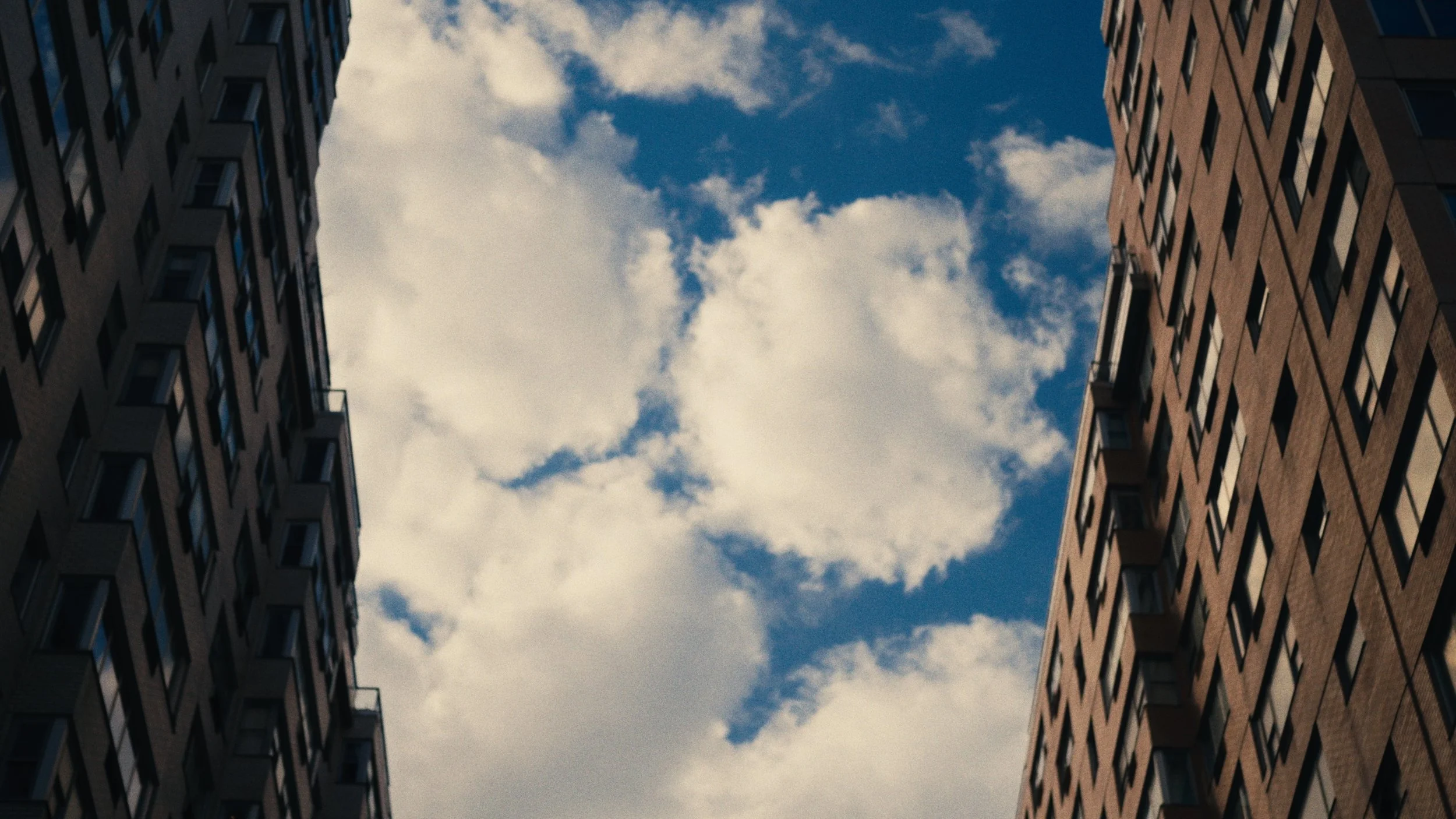 Looking up at the sky between two tall brick residential buildings with many windows and small balconies, with scattered clouds and blue sky visible.