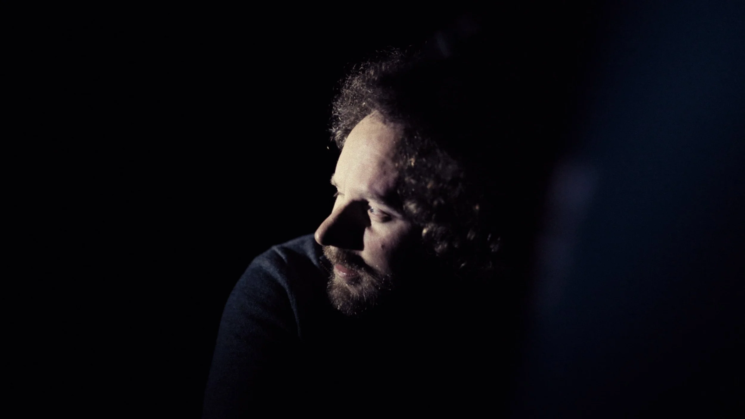Side profile of a man with curly hair and a beard, illuminated by a soft light on his face against a dark background.