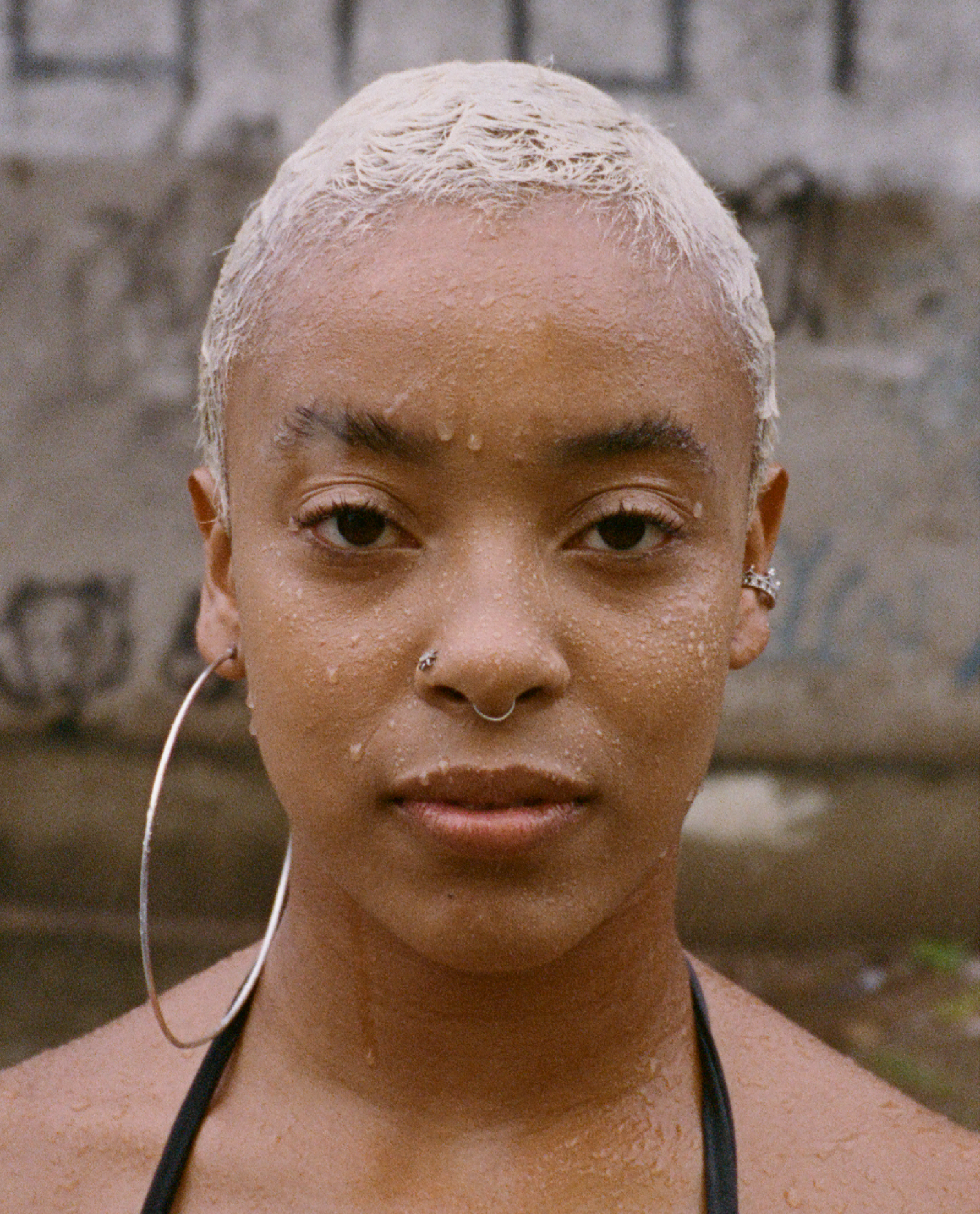 Close-up of a woman with short, platinum blonde hair, wearing multiple facial piercings, earrings, and a black top strap visible on her shoulder, standing in front of a blurred brick wall.
