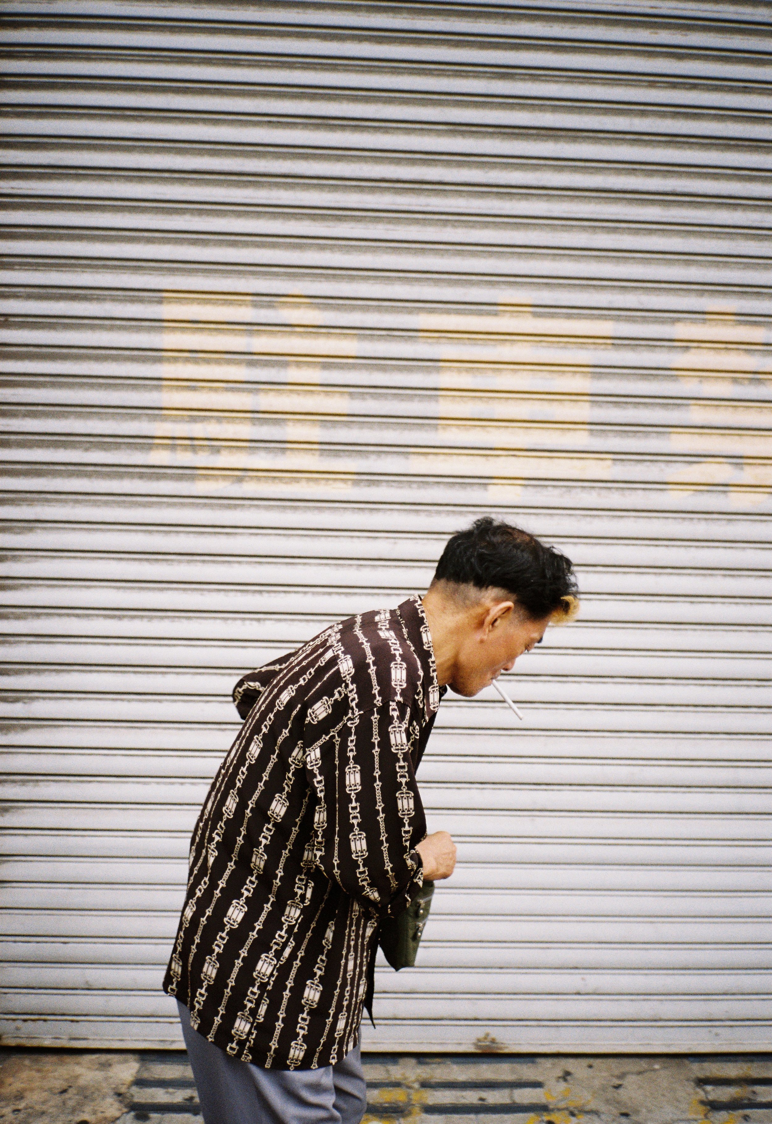 A man with a cigarette in his mouth, wearing a patterned jacket, stands in front of a closed metal shutter with graffiti or faded writing.