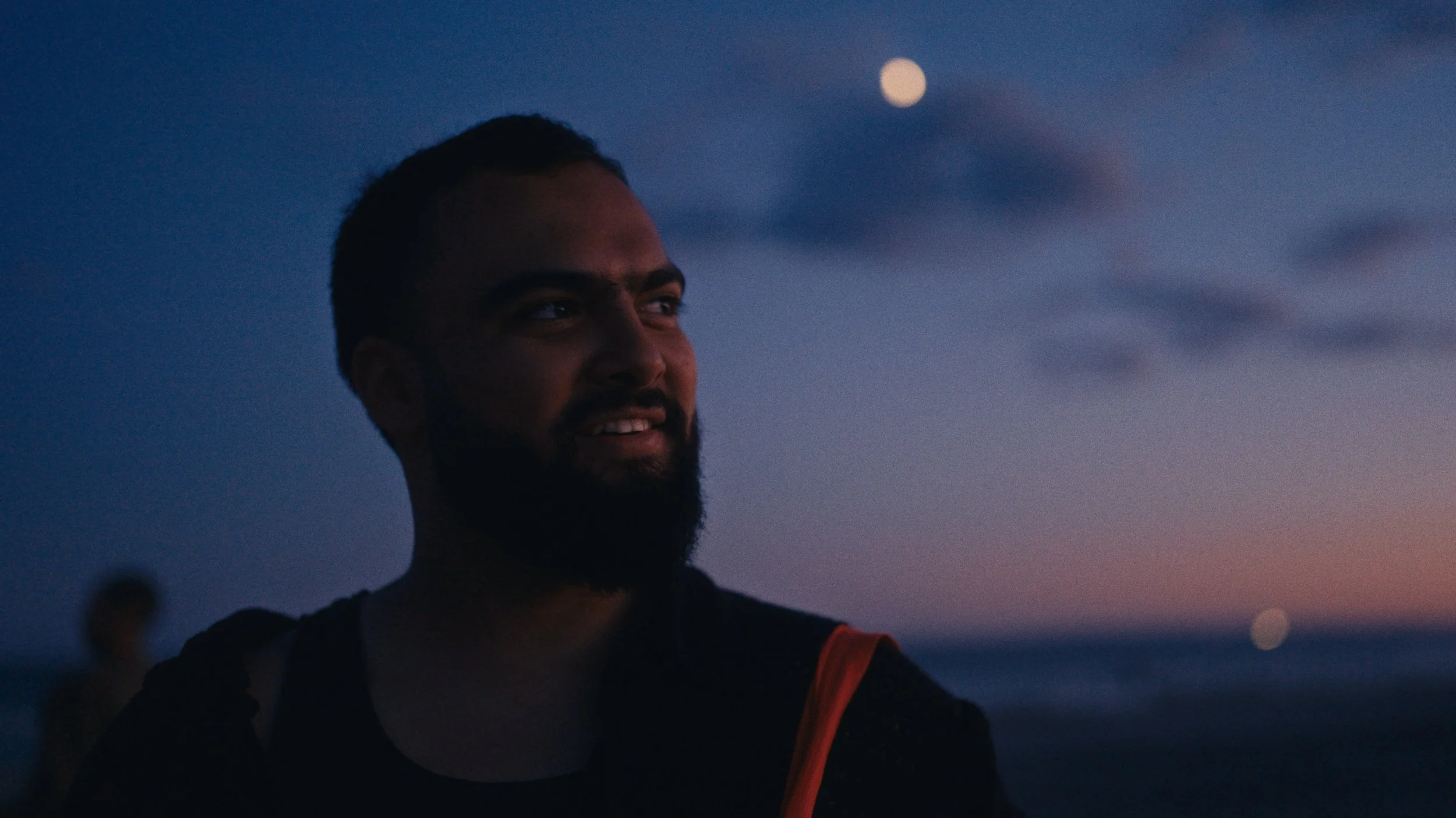 A man with a beard and short hair is smiling and looking to the side during sunset or twilight at the beach, with the moon visible in the sky.