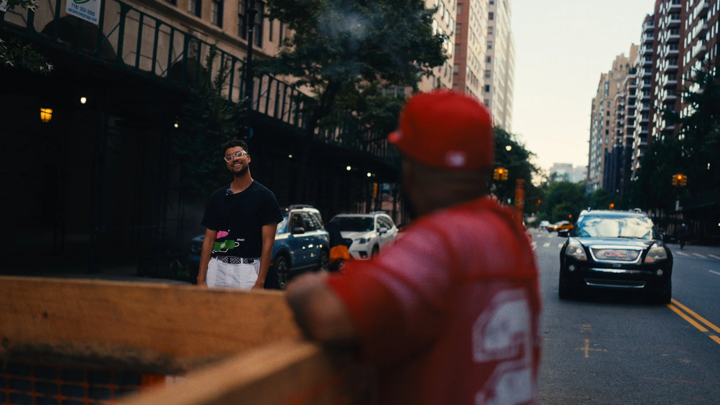 A smiling man wearing glasses and a black shirt stands on a city street, looking at a delivery person in a red cap and red shirt who is carrying a pizza box. Tall buildings and parked cars line the street in the background.