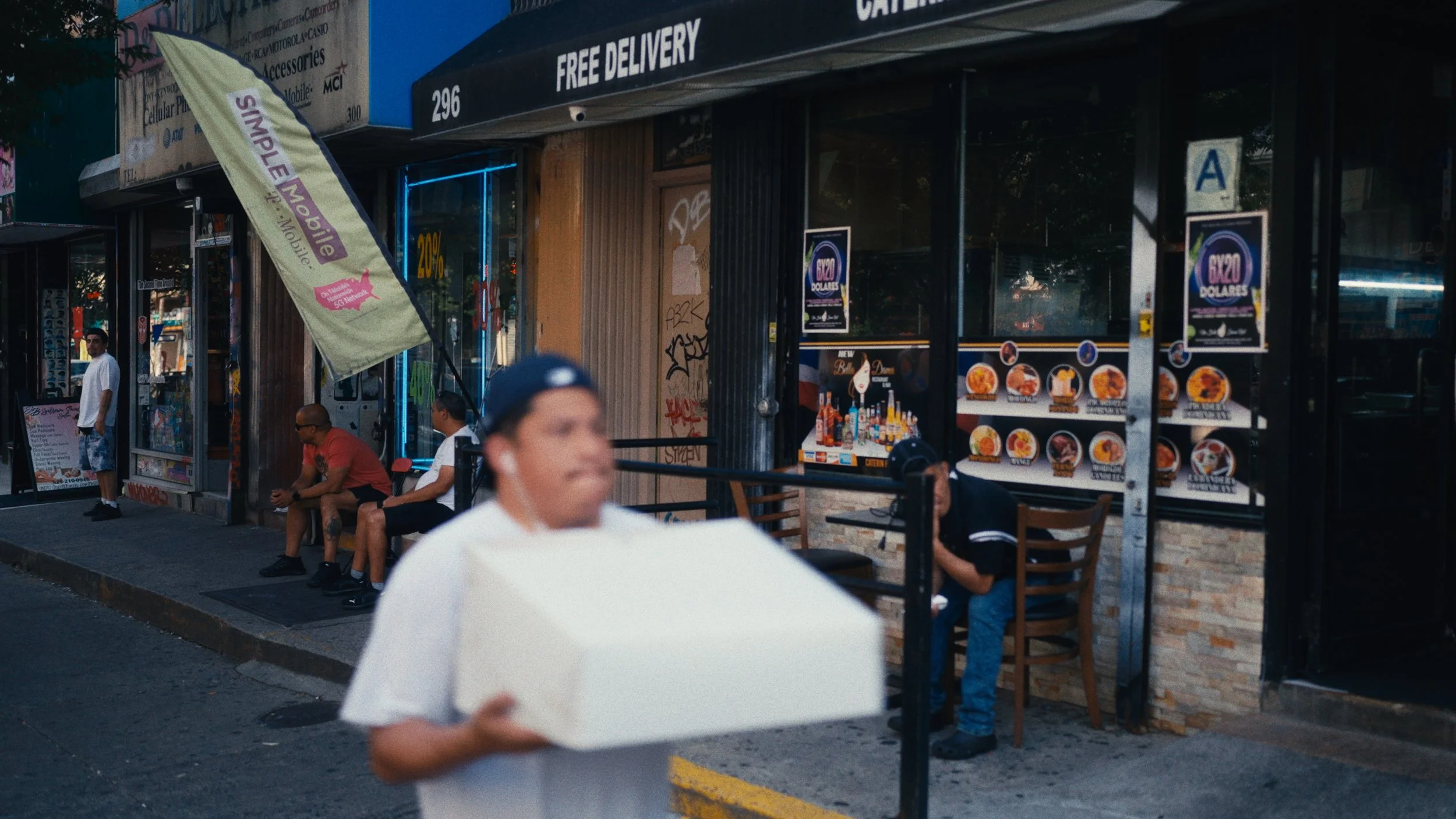 People sitting and standing outside a restaurant or food establishment on a city street, with street posters, graffiti, and a large flag advertising mobile services.