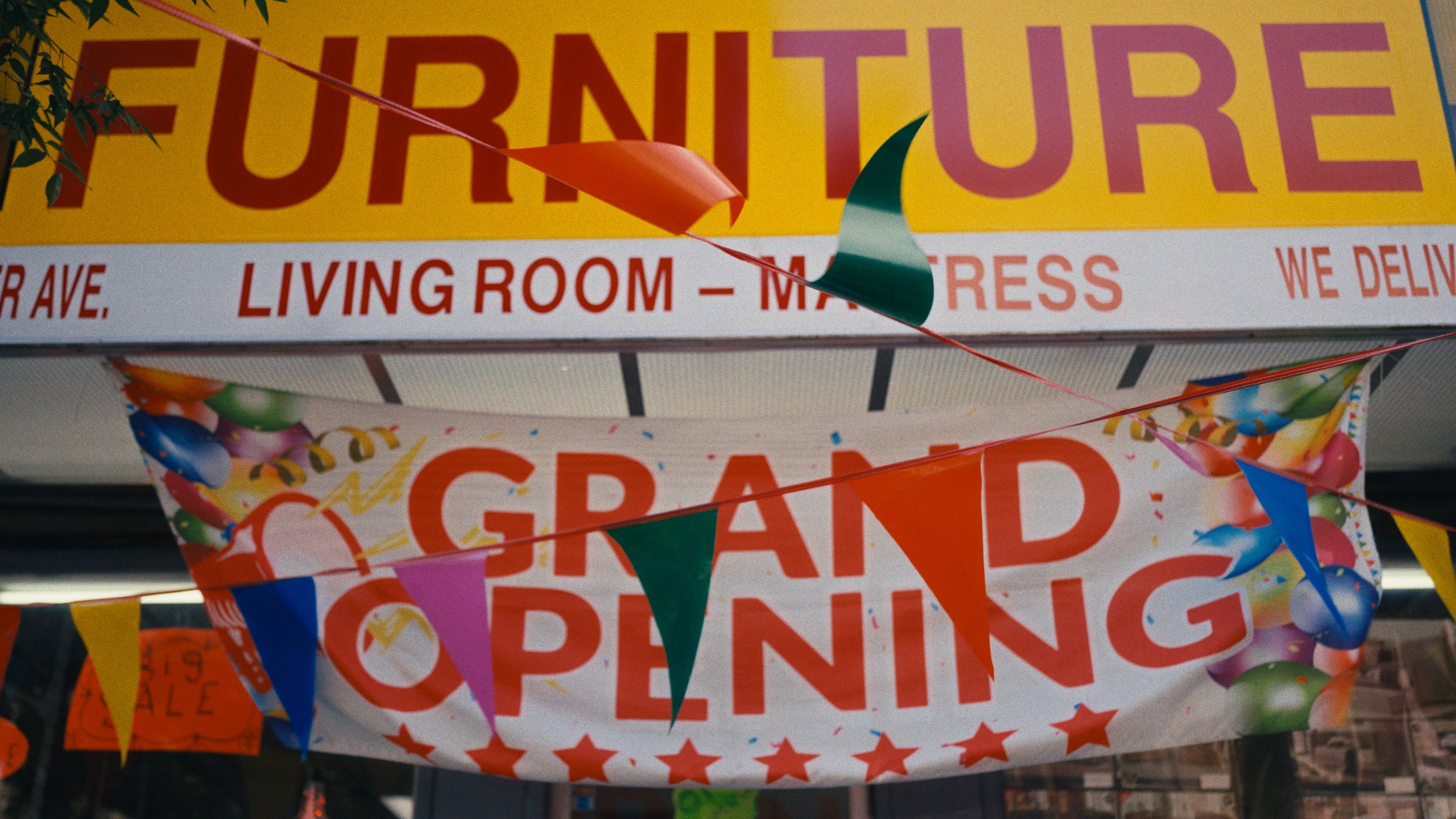 Grand opening banner with colorful balloons and pennant flags, advertising a furniture store's grand opening event.