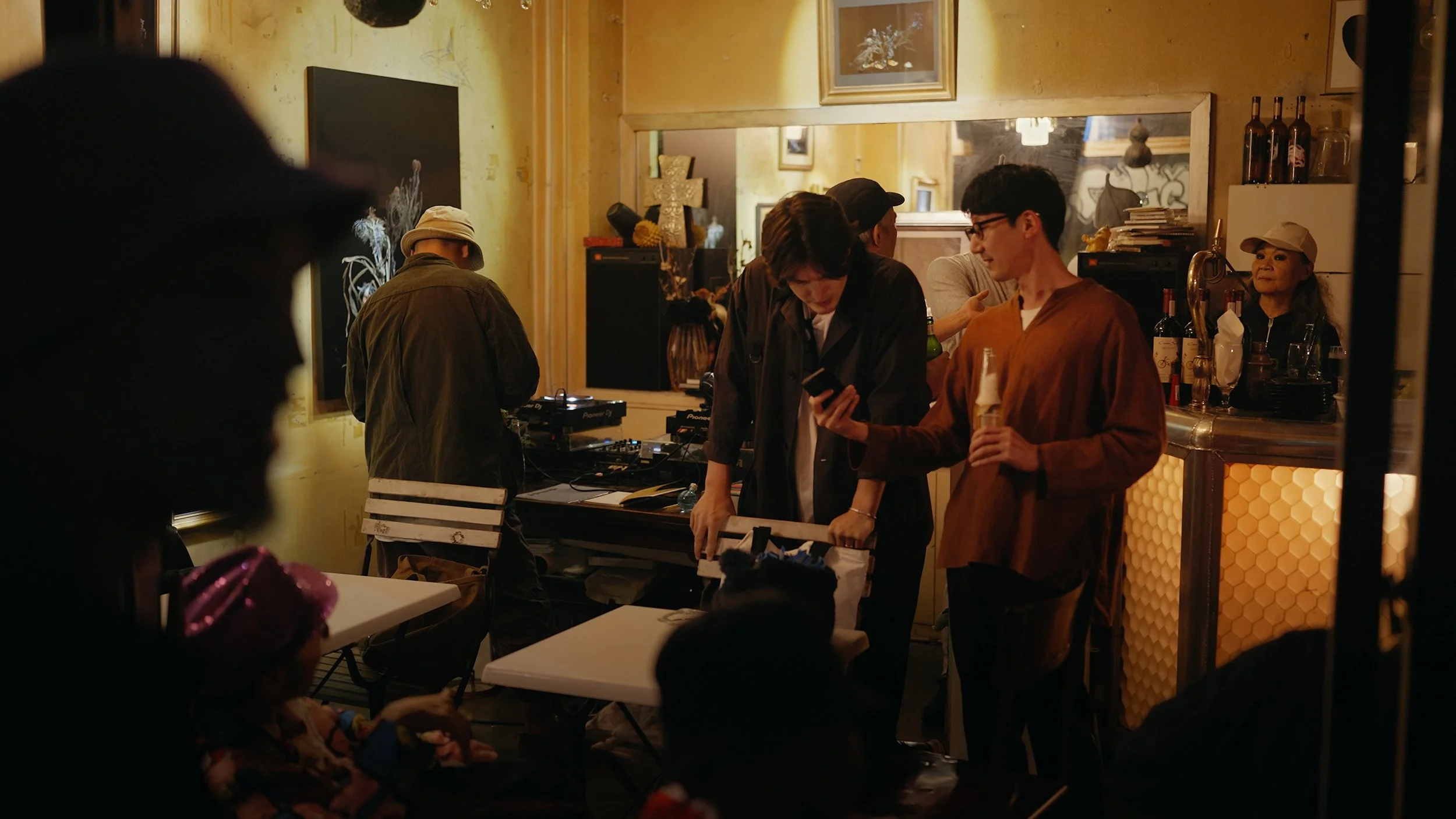 A lively indoor scene at a social gathering, where multiple people are engaged in conversations and activities near a bar area. There is a DJ setup with equipment, and the background features warm lighting and decorative objects.