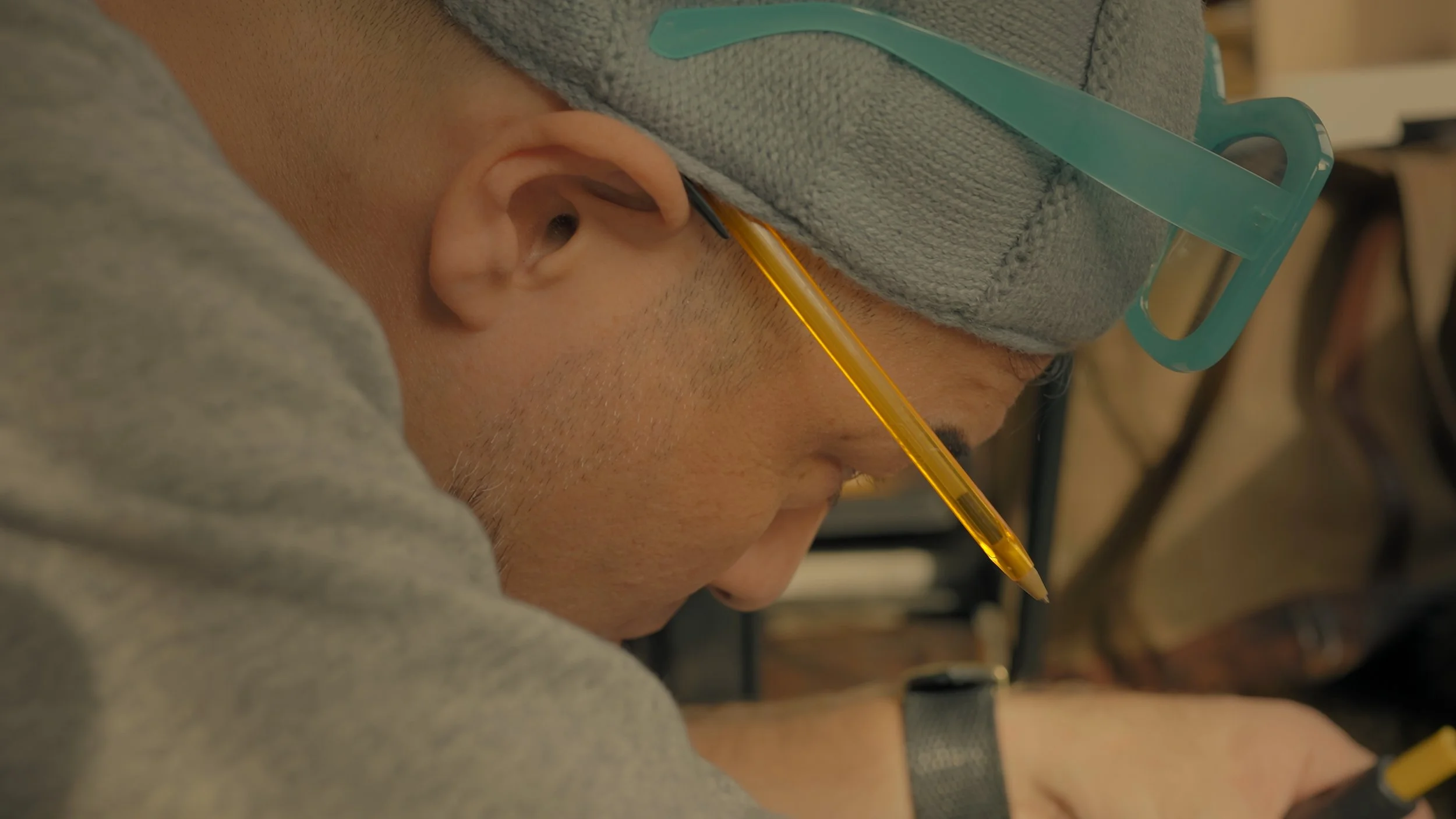 A person wearing a gray cap with teal sunglasses and a gray shirt, leaning forward with a yellow pen in their mouth, focusing on work.