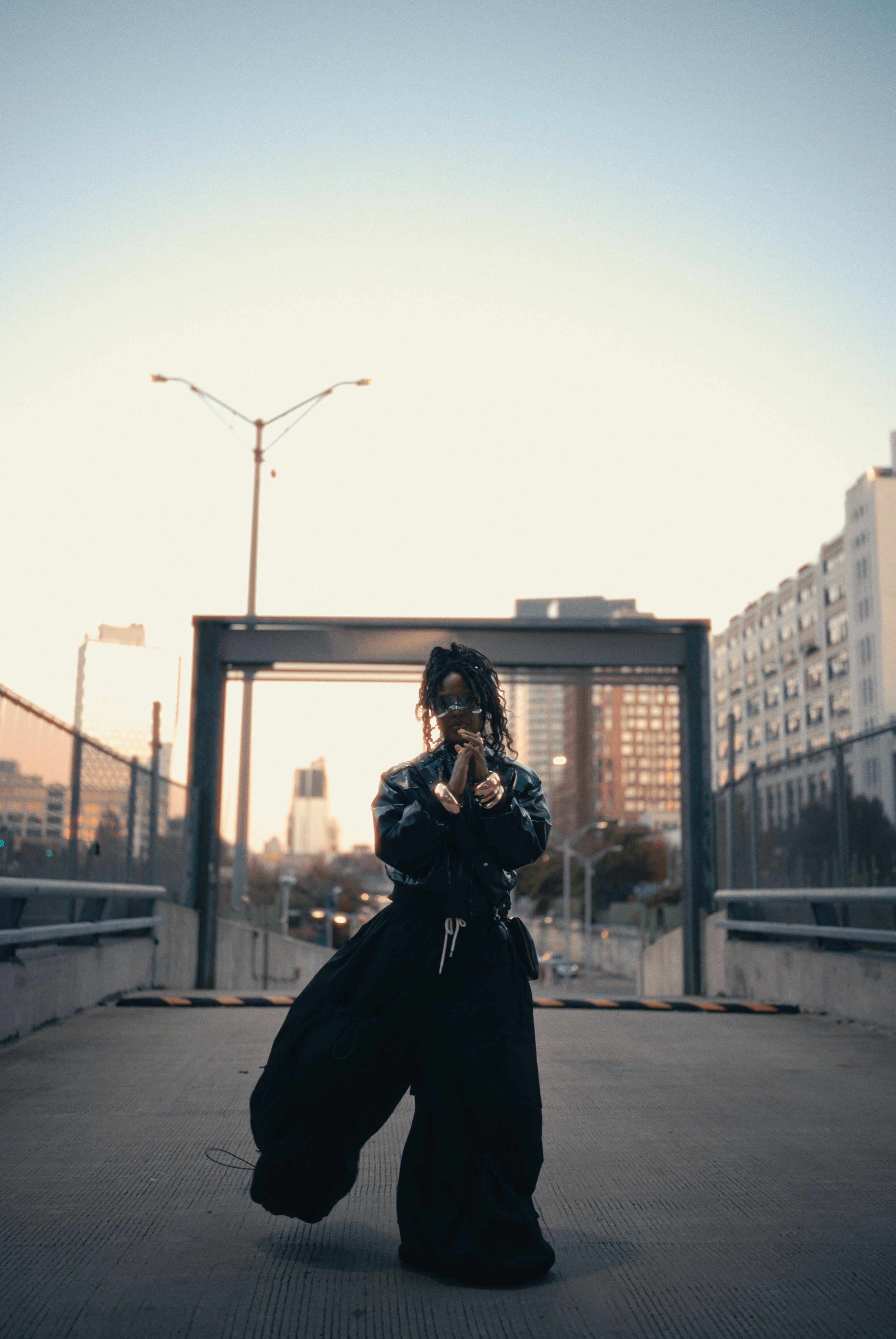 A woman standing on an urban rooftop parking lot during sunset, wearing black clothing, sunglasses, and accessories.