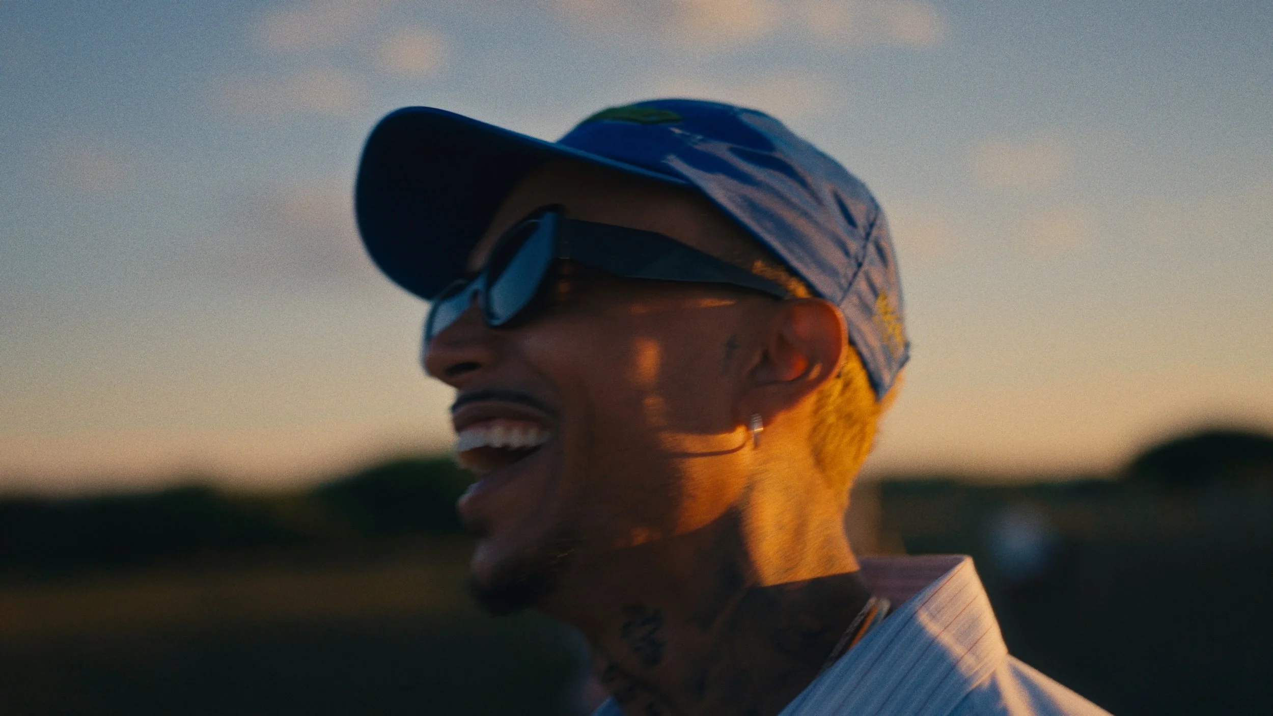A person smiling and wearing sunglasses and a baseball cap outdoors at sunset.