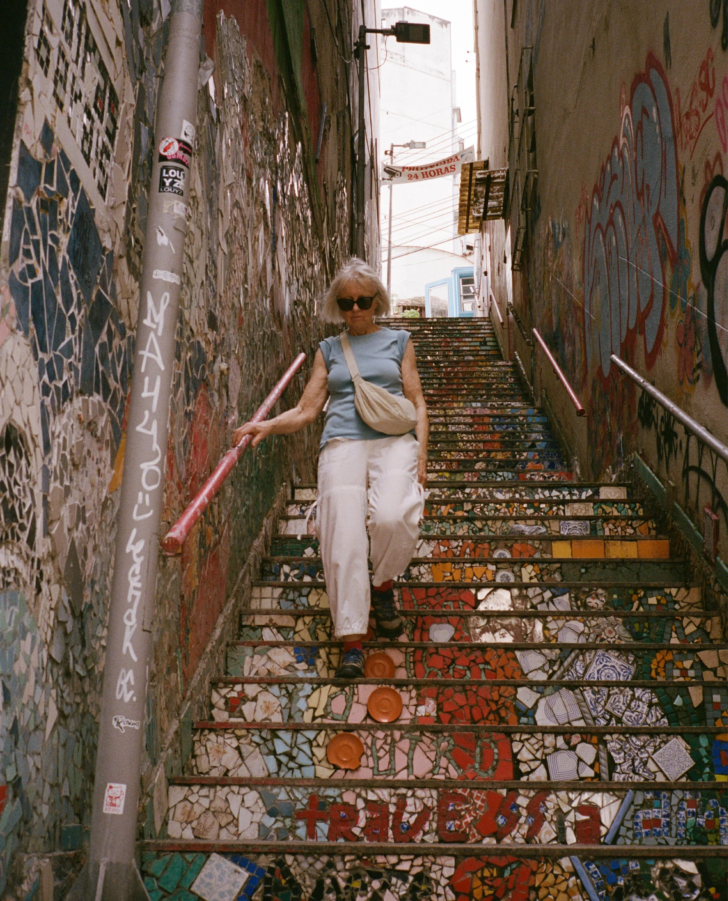 An elderly woman with blonde hair, wearing sunglasses, a light blue sleeveless top, white pants, and black shoes, is descending a colorful mosaic tiled staircase in an alleyway. She has a beige crossbody bag. The walls of the alley are covered in gra