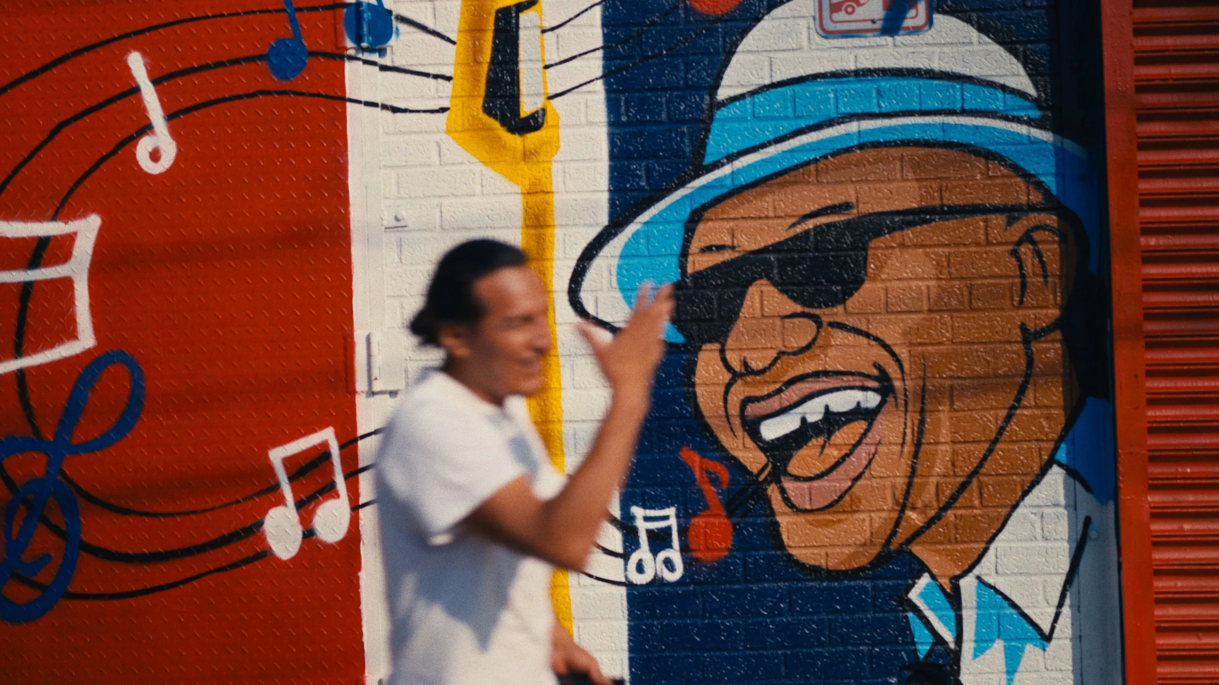 A person smiling and making a peace sign gesture in front of a colorful mural on a brick wall. The mural depicts a person with a wide smile, wearing sunglasses and a blue and white hat, with musical notes and a treble clef. The person appears blurred