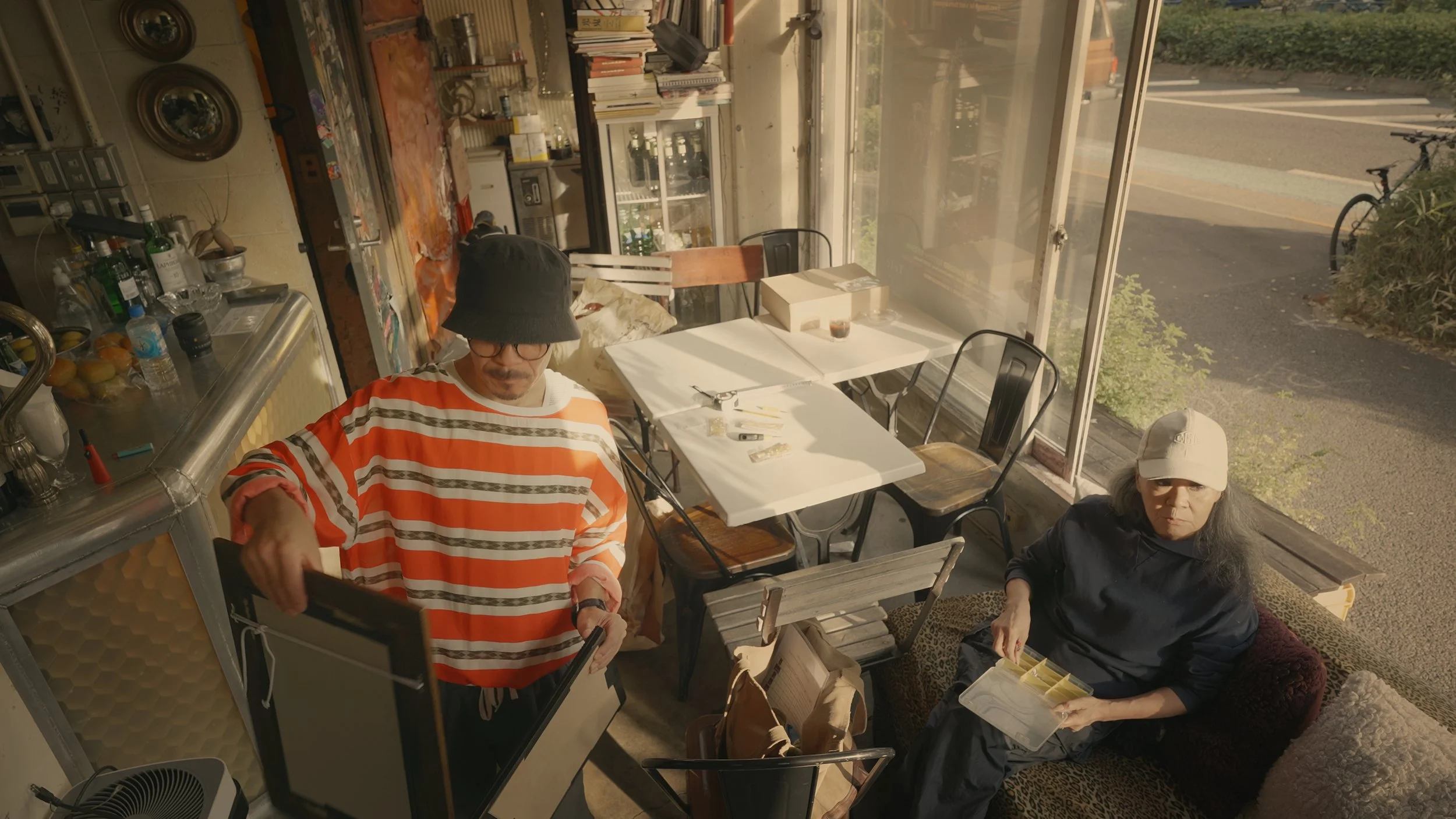 Two people sitting inside a cafe near a large window, with one person handling a tablet and the other holding a container with food, surrounded by tables, chairs, and various items on a counter, with a street visible outside.
