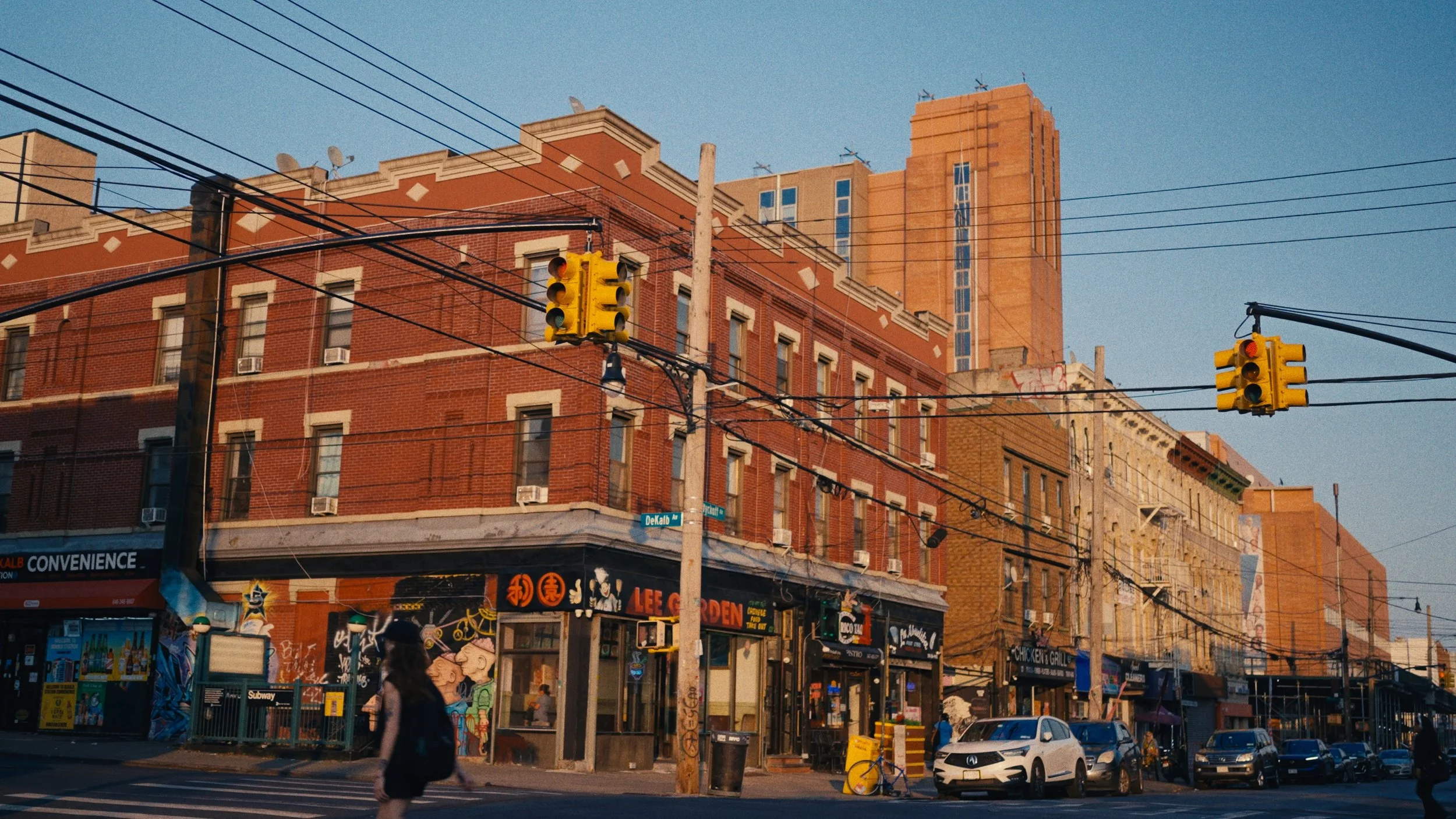 City street scene with brick buildings, traffic lights, cars parked on the street, pedestrians, and overhead power lines during daylight hours.